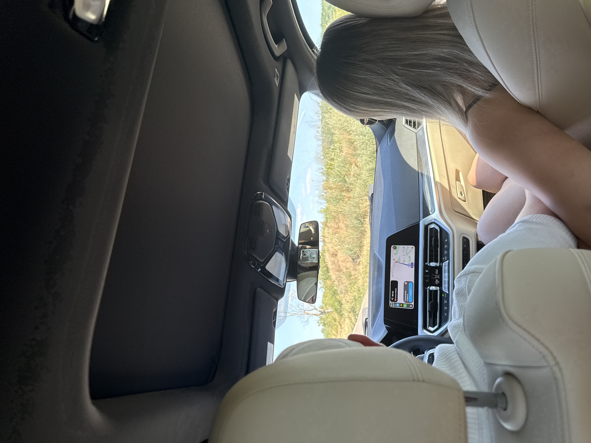 The image shows the interior of a car, viewed from the back seat, looking forward. Two people are visible: a person in the driver's seat, partially obscured by the headrest of the front passenger seat, and a person in the passenger seat, facing forward. The passenger is a woman with blonde hair, wearing a black top. The driver is wearing a white sweater. The car's dashboard features a navigation screen displaying a map and route information. Outside the car, a rural landscape with dry vegetation and a tree is visible under a clear blue sky. The car is on a road, suggesting a journey. The location is identified as Lehancea, Romania.