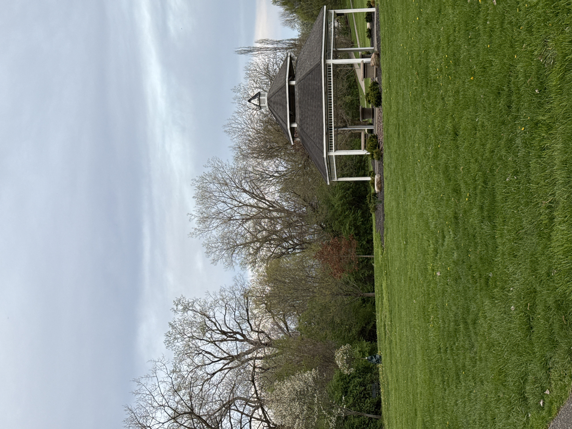 The image shows a gazebo in a grassy area. The gazebo has a dark grey roof, white supports, and is surrounded by trees. The sky is overcast. The location is likely a park in Grove City, Ohio.
