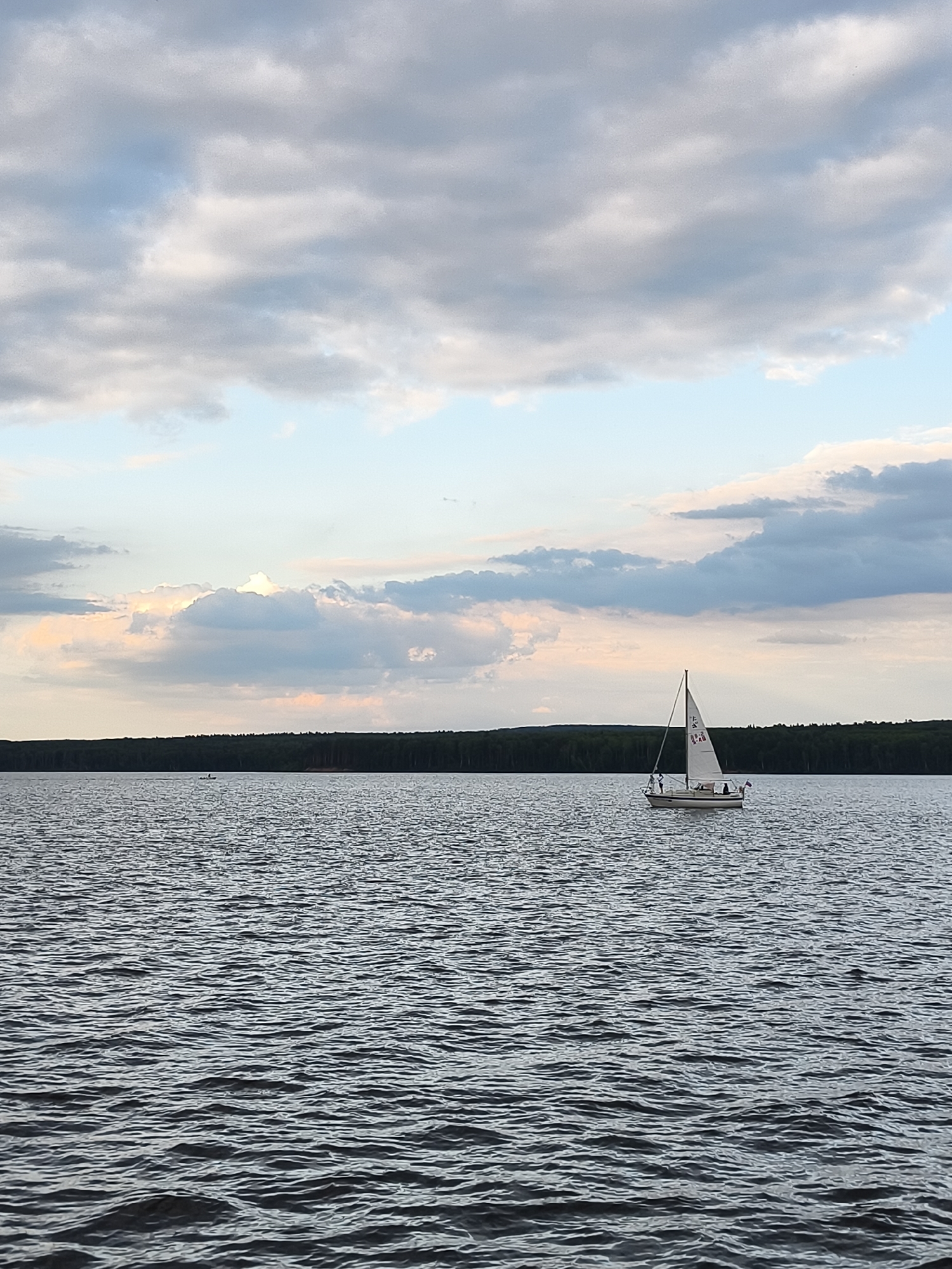 A white sailboat with a tan hull is sailing on a dark, rippling body of water. The boat has a single mast and a white sail, with some red markings on it. There appear to be a few people on the deck of the sailboat. The boat is positioned in the middle right of the frame.
In the background, a dark, dense line of trees forms the horizon, suggesting a forested area. Above the trees, a vast sky is filled with dramatic clouds. Some clouds are light and fluffy, catching the soft light, while others are darker and more textured, indicating a dynamic weather system. Patches of pale blue sky are visible between the clouds. The overall lighting suggests it is either early morning or late afternoon, with a diffused, soft light.
The scene evokes a sense of calm and serenity, with the gentle movement of the water and the solitary sailboat under the expansive sky. The location appears to be a large lake or a wide river, likely in a natural, somewhat remote setting, consistent with the Chaikovsky, Russia context. There is no visible text, other than some small markings on the sail of the sailboat.