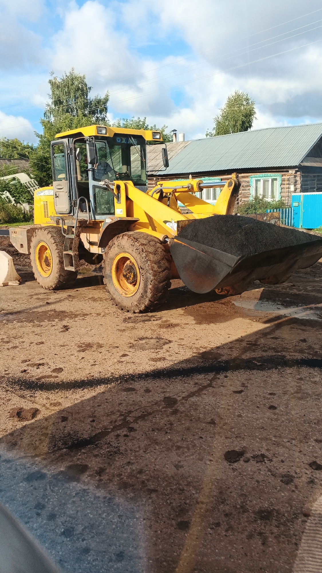 A yellow front-end loader is parked on a dirt and gravel surface. The loader's bucket is filled with dark material, possibly asphalt or soil. The machine has large, muddy tires. In the background, a wooden house with a green metal roof and a blue fence can be seen. Trees are visible behind the house, and the sky is partly cloudy. The scene appears to be in a rural or suburban area in Киясово, Russia, likely a construction or road work site.
