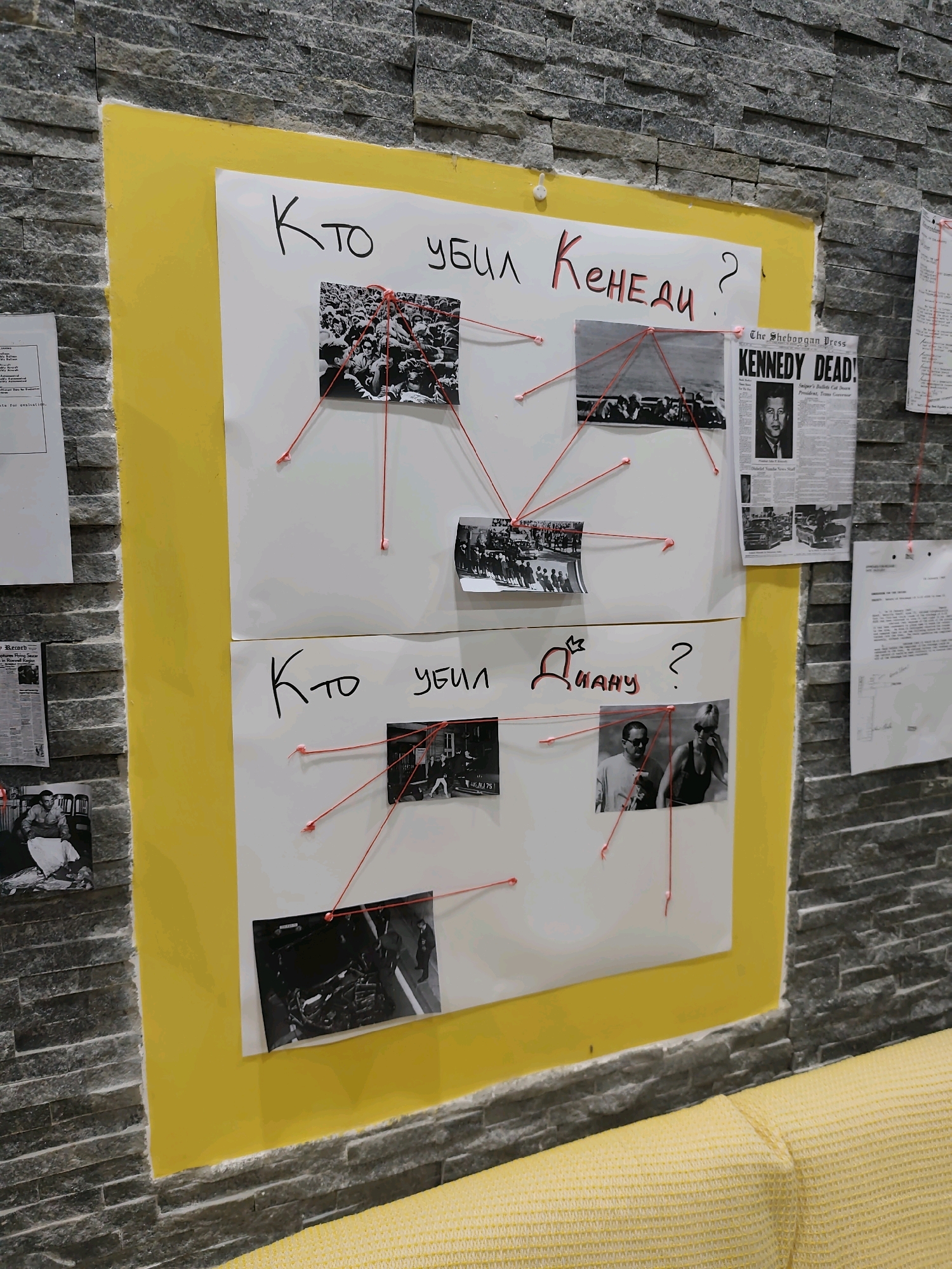 This is a forensic, matter-of-fact description of a scene that appears to be a conspiracy board, likely in a private residence or office space, in the city of Unknown, Unknown. The board is mounted on a textured, gray stone wall, with a bright yellow border. The board itself is composed of two large white sheets of paper, attached one above the other, with red string connecting various photographs and documents.
The top sheet of paper has the Russian text "Кто убил Кененди?" (Who killed Kennedy?) written in black marker. This sheet contains three photographs and a newspaper clipping. The first photograph, to the left, shows a crowd of people. The second photograph, in the center, depicts a motorcade, likely in Dallas. The third photograph, to the right, is a newspaper headline reading "KENNEDY DEAD!". Red strings connect these items, forming a web of connections.
The bottom sheet of paper has the Russian text "Кто убил Диану?" (Who killed Diana?) written in black marker, with a crown drawn above the letter "и" in "Диану". This sheet also contains three photographs. The photograph on the left shows a man seated in a chair, wrapped in a white cloth or sheet. The central photograph shows two individuals, a man and a woman, the woman wearing sunglasses. The photograph on the right shows what appears to be a scene of investigation or recovery, with debris and several people in the background. Red strings connect these photographs.
Other documents are partially visible on the right side of the image, also connected by red string to the main board. These appear to be official-looking papers, some with text that is illegible in this image. A yellow upholstered surface, possibly a sofa or chair, is visible in the lower part of the frame. The overall impression is that of a detailed investigation or a display of theories regarding the assassinations of John F. Kennedy and Princess Diana.