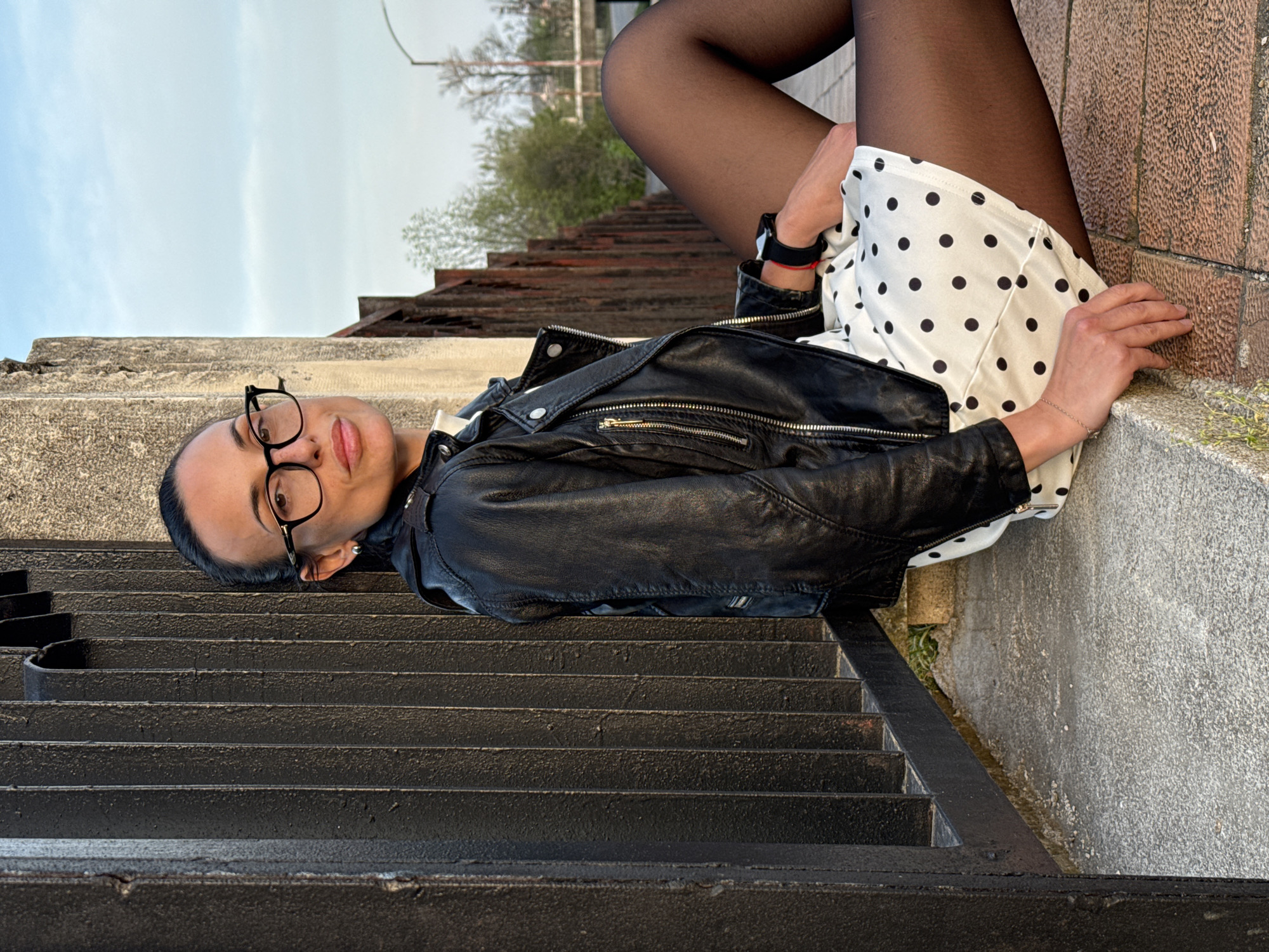 A young woman is captured in a full-body shot, lying on concrete steps or a ledge in an outdoor setting. She is oriented horizontally in the frame, leaning her upper body against a textured wall made of brick or rough stone. Her head is slightly elevated, and she gazes directly at the camera with a calm, subtle smile, wearing black-framed glasses.
She is dressed in a black leather jacket, a white dress or skirt adorned with black polka dots, and black sheer tights or stockings. Her accessories include a black watch or bracelet and a red string bracelet on her left wrist, and a delicate silver chain bracelet on her right wrist. Her dark hair is pulled back, creating a neat silhouette against her face.
Below her (to the viewer's left in the rotated image), a set of dark, possibly metal, stairs descends, leading away from the immediate foreground. The background features a light blue sky with some cloud cover, and distant blurred trees or foliage, hinting at a natural element within an urban or semi-urban environment. The lighting is soft and natural, suggesting it is either late afternoon or early evening.
The scene conveys a relaxed atmosphere, with the woman appearing comfortable and at ease. There is no visible text in the image. This outdoor setting, with its architectural elements and natural backdrop, is located in Pleven, Bulgaria.