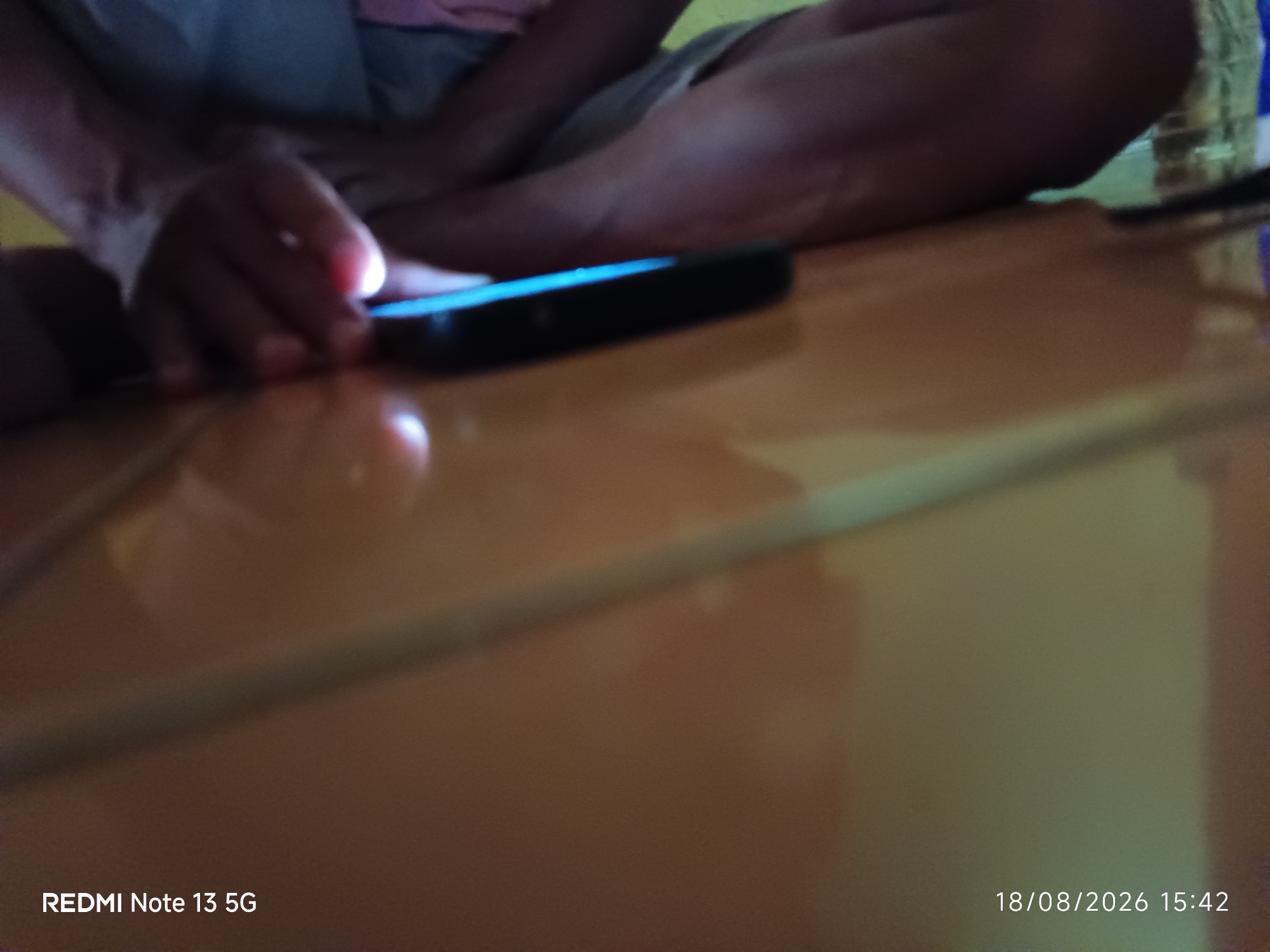 The image shows a close-up of a person's hands holding a smartphone. The phone is black and its screen is illuminated with a bright blue light. The person appears to be sitting down, with their legs visible in the background. The overall lighting is dim, and the focus is on the phone and hands. The lower right corner of the image contains a date and time stamp: "18/08/2026 15:42". The lower left corner has text that reads "REDMI Note 13 5G". The scene is set on a reflective orange surface. There is no specific information in the image to determine the location within Talaga Sari, Indonesia.