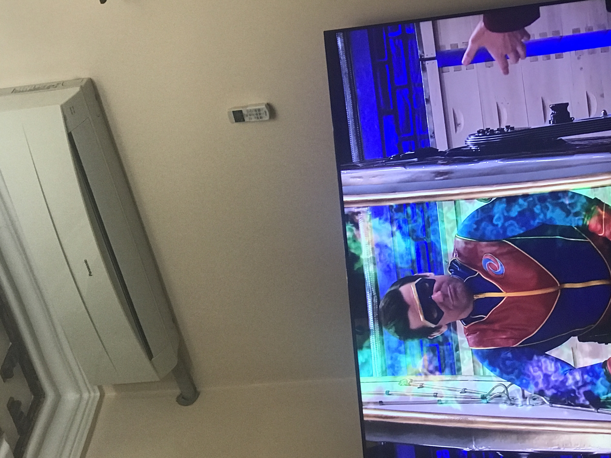 A medium, color, slightly rotated, indoor photograph depicts a television on a wall adjacent to an air conditioner. The television shows a scene from a program with a person in a blue and maroon outfit with a blue patterned design on the legs, wearing sunglasses. The person appears to be lying on a surface. Above this scene, another scene is visible, showing a white surface with blue stripes and a hand reaching down. To the left of the television, an air conditioner is mounted on the wall, angled slightly downwards. Above the air conditioner and to the right, a small white remote control is affixed to the wall. The environment appears to be a domestic setting, possibly a living room or bedroom. There is no indication of the location being Yerevan, Armenia.