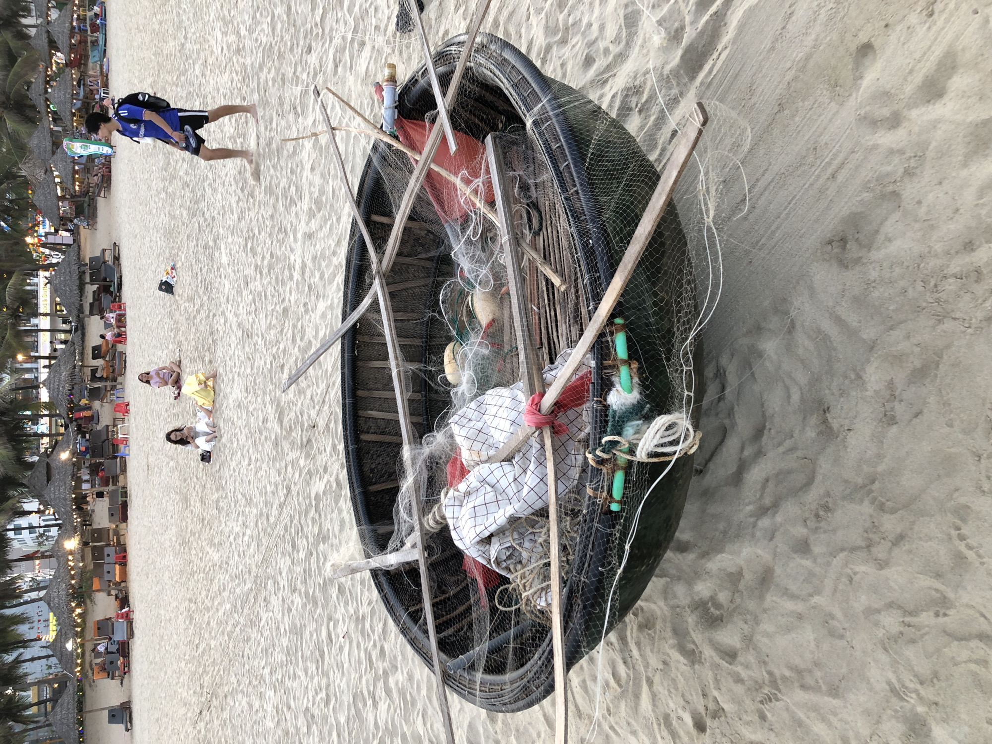 This image captures a busy beach scene in Đà Nẵng, Vietnam. The foreground is dominated by a traditional Vietnamese coracle boat, filled with fishing nets, ropes, and some white fabric. Several wooden poles are propped up around the boat, likely to support the nets or for fishing purposes. The boat rests on a sandy beach, with ripples in the sand suggesting it was recently placed there or disturbed by activity.
In the background, a row of thatched umbrellas and beach chairs creates a resort-like atmosphere. Lights are strung between some of the umbrellas, indicating that it might be late afternoon or early evening. Several people are visible in the distance, some sitting on the beach and others in the seating areas under the umbrellas, suggesting a leisurely day at the beach.
A young man wearing a blue shirt, black shorts, and a backpack is walking on the sand, facing away from the camera. Further down the beach, three women are sitting on the sand. One is wearing a yellow dress, another a pink top, and the third is in white clothing. They appear to be casually interacting or relaxing. The overall mood of the image is relaxed and vibrant, typical of a popular tourist beach. The lighting suggests it is not the middle of the day, possibly late afternoon or early evening.