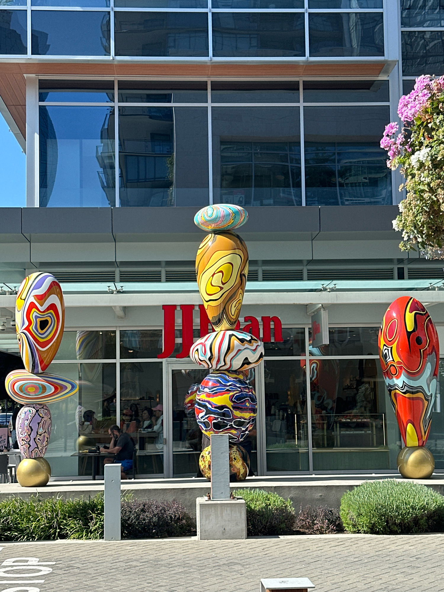 This image depicts the exterior of a retail establishment, identified by prominent red lettering spelling "JIFFAN" above the storefront windows. The building features a modern architectural design with large, reflective windows and a light-colored facade. In front of the building, a series of colorful, abstract sculptures are displayed. These sculptures are stacked and appear to be made of a glossy material, with swirling patterns and bright, varied colors. Some of the sculptures are oval or teardrop-shaped, while others are more rounded. In the foreground, a man is seated at a table outside the store, appearing to be engaged with a tablet or book. The area in front of the sculptures and the store is paved, and there is a small patch of green landscaping. The sky above is clear and blue. This scene is located in Burnaby, Canada.