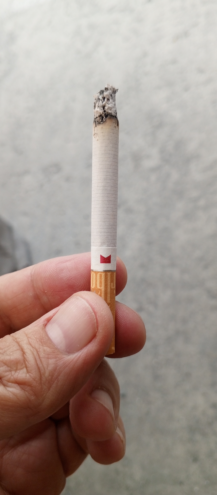 In Yeruda, Russia, a person's hand is holding a lit cigarette. The cigarette has a white paper wrapper with a red and white logo near the filter. The tip of the cigarette is burning, and ash is visible. The background is a blurry, light gray concrete surface.