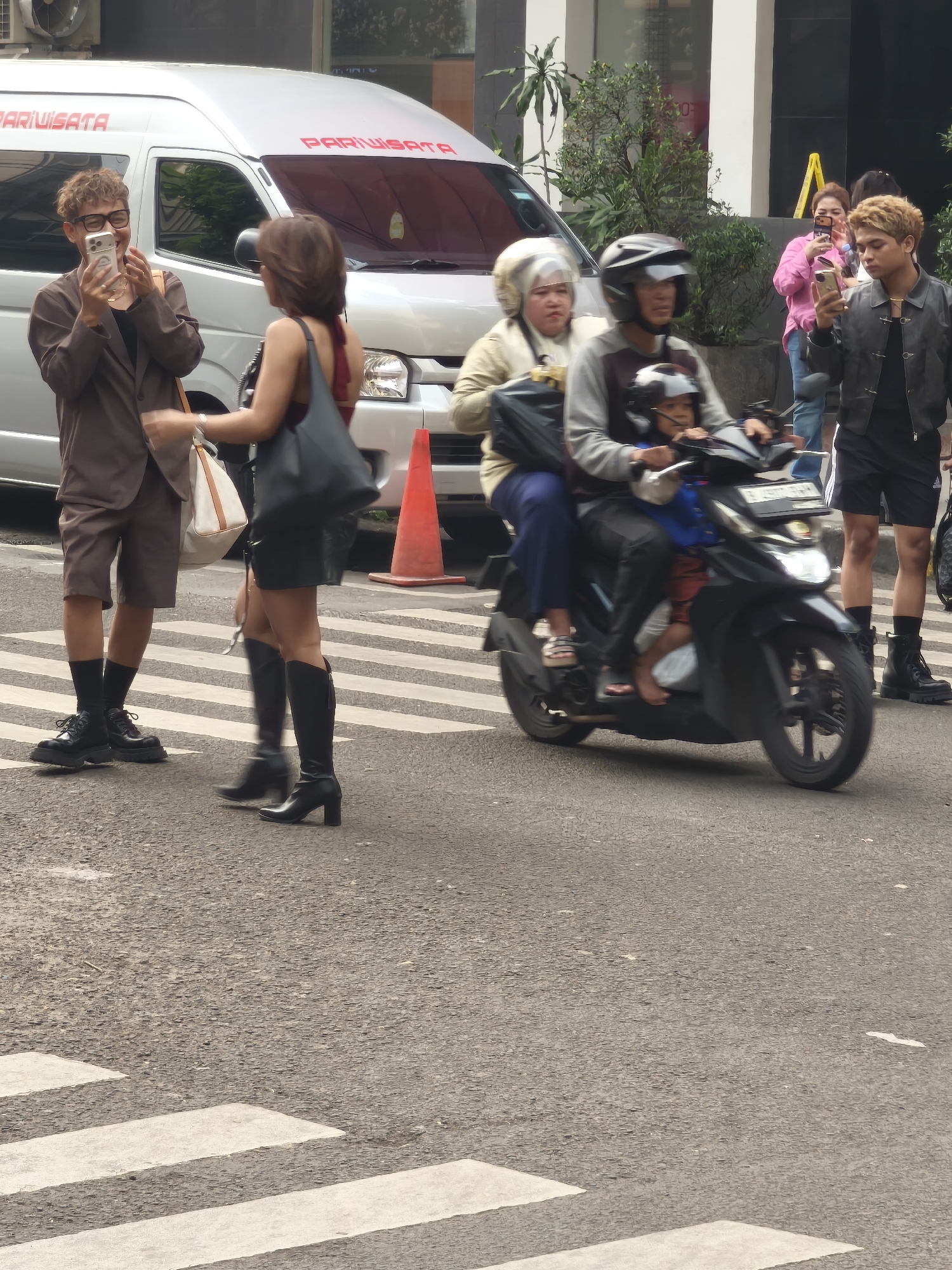 On a city street in South Jakarta, Indonesia, a group of individuals are captured in a scene that appears to be near a pedestrian crossing. In the foreground, a man with curly hair and glasses, wearing a brown suit jacket and shorts, is holding up a smartphone to take a photo or video. He is standing on a pedestrian crossing with white stripes. Beside him, a woman with dark hair, wearing a dark top and a short skirt, is walking away from the camera, carrying a dark bag over her shoulder. She is also on the pedestrian crossing and is wearing black boots.
In the mid-ground, a motorcycle with three passengers is moving from left to right across the scene. The driver, wearing a helmet, is a man. A woman in a light-colored jacket and headscarf is seated behind him, and a child wearing a helmet is seated in front of the driver. Behind them, another man with blonde hair, wearing a black leather jacket and shorts, is looking at his smartphone. Further in the background, a woman in a pink shirt and blue jeans is holding up her smartphone. A white van with the word "PARIWISATA" (Indonesian for tourism) written in red on its side is parked in the background. An orange traffic cone is placed on the side of the road. The environment suggests a busy urban setting with buildings visible in the background.