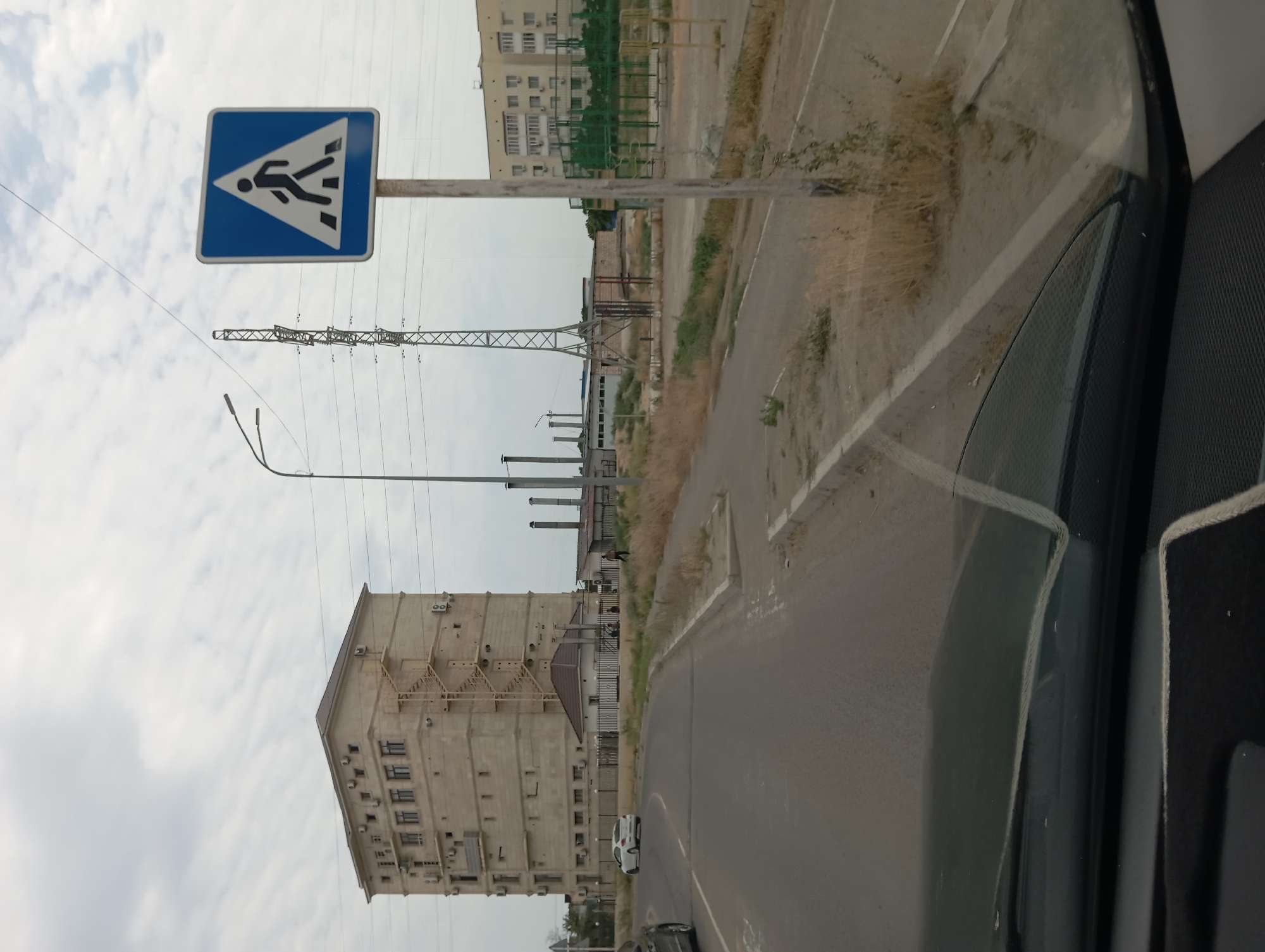 The image captures a street scene in Aktau, Kazakhstan, on an overcast day. A multi-story concrete building with numerous air conditioning units and an external metal staircase is prominent on the left. A white sedan is visible on the road in front of the building. To the right of the road, a pedestrian crossing sign with a silhouette of a person walking is mounted on a pole. Beyond this, industrial structures, including a tall, lattice metal tower and several low-lying buildings with smokestacks, are present. In the far right of the image, a light-colored residential building with multiple windows and balconies is visible, adjacent to an enclosed outdoor sports court with green fencing. The foreground shows the interior of a car, with the windshield and dashboard visible. The environment is arid, with dry grass and sparse vegetation lining the road and surrounding the buildings.