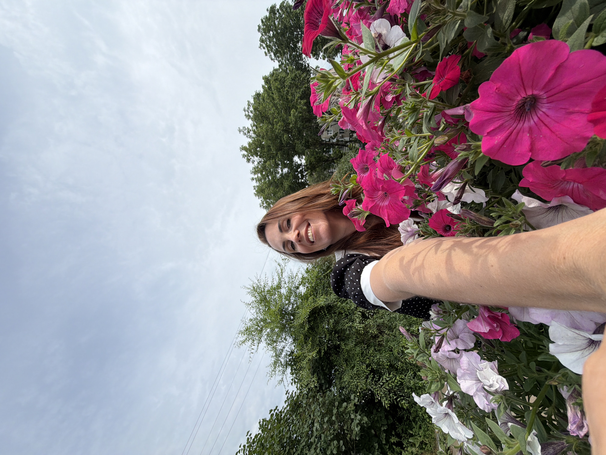 A first-person perspective selfie is captured from a low angle, showcasing a woman in the foreground amidst a vibrant display of pink and white petunia flowers. The woman, with long, reddish-brown hair and a bright smile, is looking directly at the camera. She is wearing a black and white polka-dot glove on her right hand, which is extended towards the camera, likely holding the phone for the selfie. Her left arm is partially visible. Behind the flowers, lush green trees and foliage form a backdrop, with a pale, cloudy sky overhead. The scene appears to be outdoors, possibly in a garden or park. The presence of telephone wires in the upper left quadrant suggests an urban or suburban setting. No specific identifying landmarks or architectural features are discernible to pinpoint the exact location within Birobidzhan, Russia, but the overall setting is consistent with an outdoor, green space within the city.