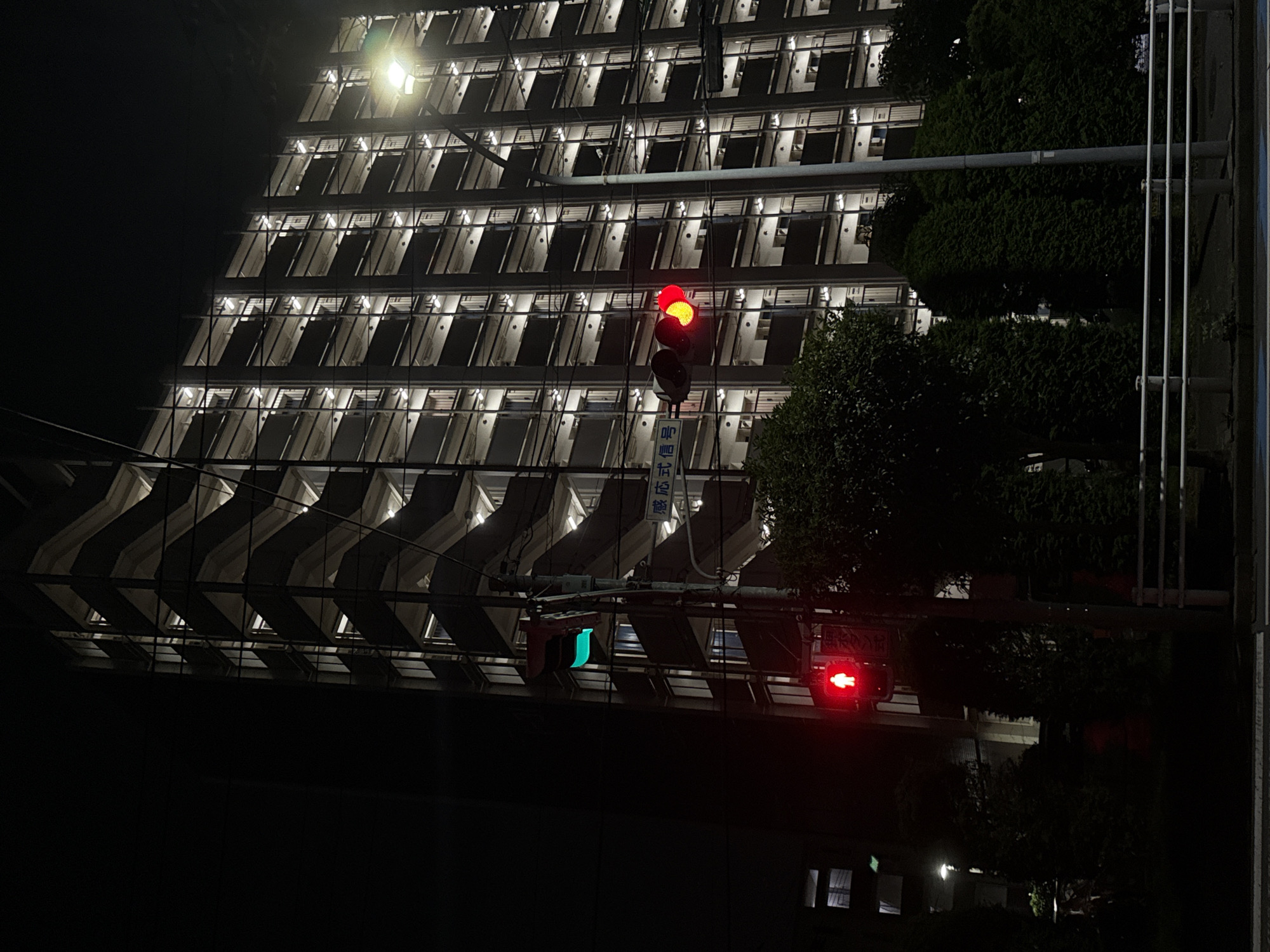 The image shows a street view at night in Yachiyo, Japan, focusing on a tall, multi-story apartment building with numerous balconies and exterior staircases illuminated from within. A traffic light is prominently featured in the foreground, displaying a red light for pedestrians. To the left of the pedestrian signal, a traffic signal shows a green light. The building is dark except for the warm interior lighting visible through the windows. Trees and foliage are visible at the base of the building and to the right of the traffic light. A white metal railing runs along the edge of the street in the lower part of the image. The sky is dark, indicating nighttime.