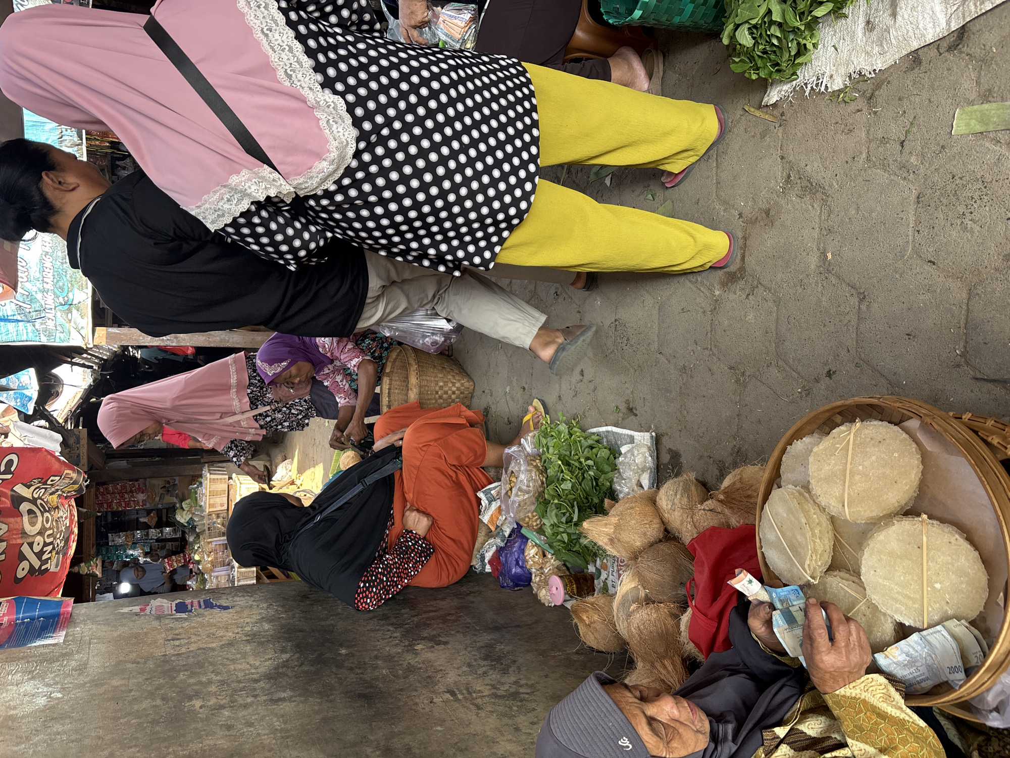 The image depicts a bustling street market scene in Tirtomoyo, Indonesia. In the foreground, an elderly woman wearing a grey hijab and a patterned gold jacket is counting money in front of a woven basket filled with round, flat crackers. To her right, a pile of coconuts is visible. Behind her, a woman in an orange skirt and black hijab is seated, her hands resting on her lap. Further into the market, a person in a pink hijab and a black and white polka dot top is standing, their back to the camera, and is wearing bright yellow trousers. Another person in a black shirt and light-colored pants is walking away. Several stalls are visible in the background, displaying various goods. The ground is paved with hexagonal stones, and the overall atmosphere suggests a lively marketplace.