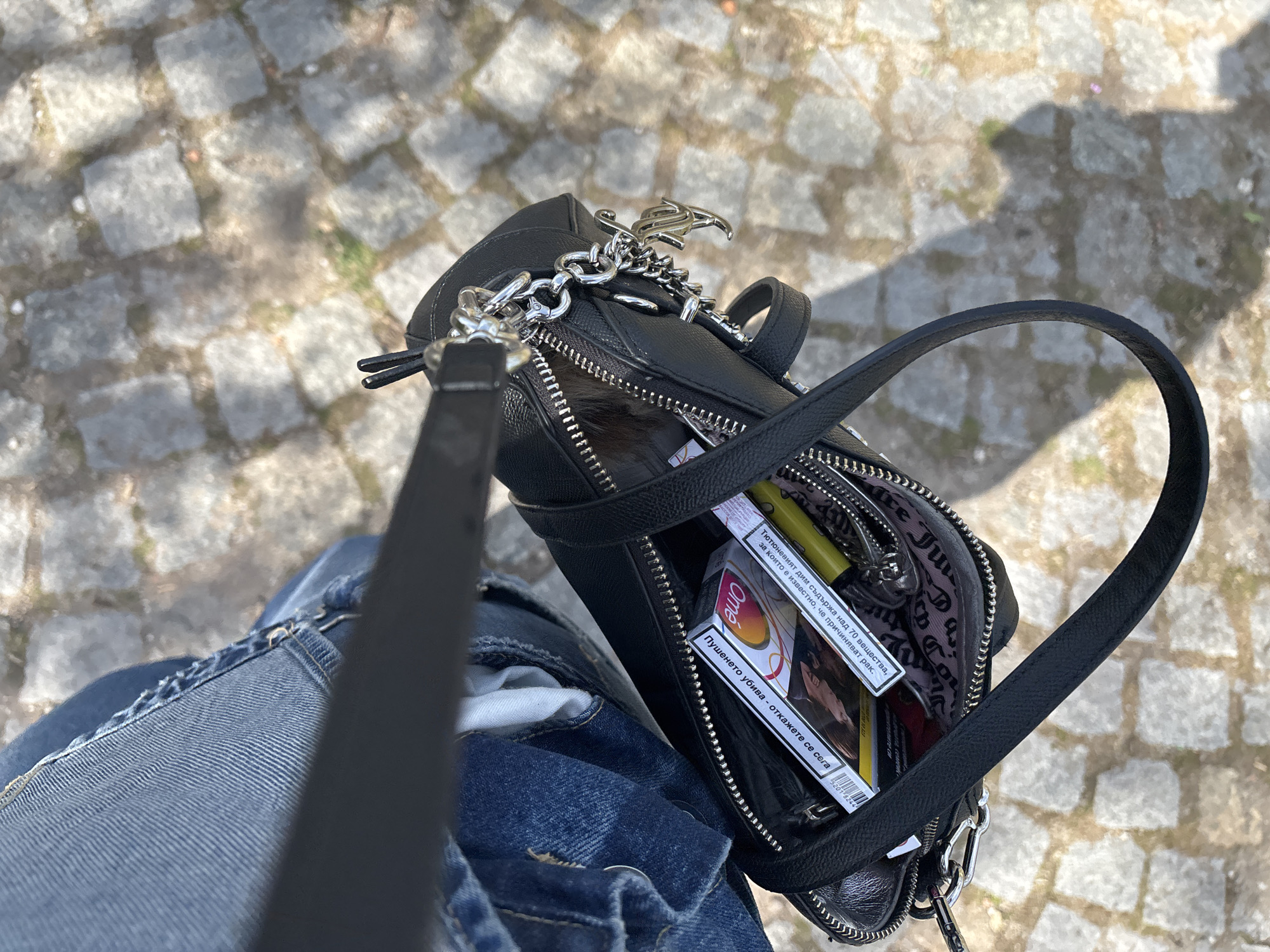 A close-up, high-angle shot captures the contents of an open black leather handbag, held by an individual wearing denim jeans, while standing on a cobblestone surface. The bright sunlight suggests daytime and clear weather, casting distinct shadows on the ground.
Inside the open handbag, a pack of "Omé" cigarettes is clearly visible, along with a yellow lighter. The lining of the bag features a pattern with the "Juicy Couture" logo, which is also present on a metallic charm attached to the bag's chain.
The visible text on the cigarette pack is in Bulgarian, reading: "Тютюневият дим съдържа над 70 вещества, за които е известно, че причиняват рак." (Tobacco smoke contains over 70 substances known to cause cancer.) and "Пушенето убива - откажете се сега" (Smoking kills - quit now). Part of a number "5201" is also visible, likely from a barcode.
The scene appears to be an everyday moment outdoors, possibly in Plovdiv, Bulgaria, given the Bulgarian text on the cigarette packaging. No specific activity beyond holding the bag is depicted.
