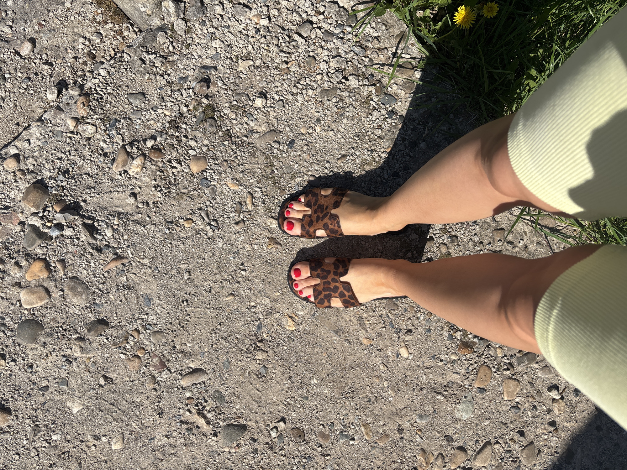 A high-angle photograph shows a person's legs and feet in leopard-print sandals standing on a gravelly surface with patches of grass at the upper right corner. The person is wearing light green clothing. The toenails are painted red. The ground is composed of small rocks and dirt, suggesting a path or a roadside area. The lighting is bright and sunny, casting shadows on the ground. The location is assumed to be in the city of Ферма, Russia.