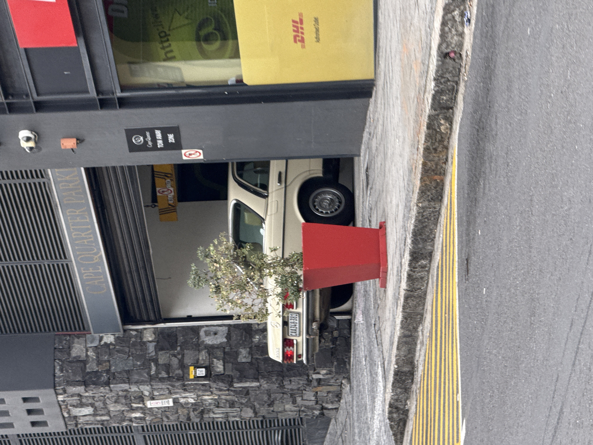 The image captures a scene at the Cape Quarter Park in Cape Town, South Africa. An older model, light-colored car with license plate "CAA348-381" is partially parked under a building overhang. The building has visible signage for "Cape Quarter Park," "Cape Quarter Tow Away Zone," and "DHL." A red, rectangular planter is positioned on the sidewalk next to the car. The sidewalk has yellow and black striped markings, and the street is made of asphalt. The building's exterior is constructed with dark stone blocks, and there's a small tree or bush visible beside the car. A surveillance camera is attached above the entrance of the building.