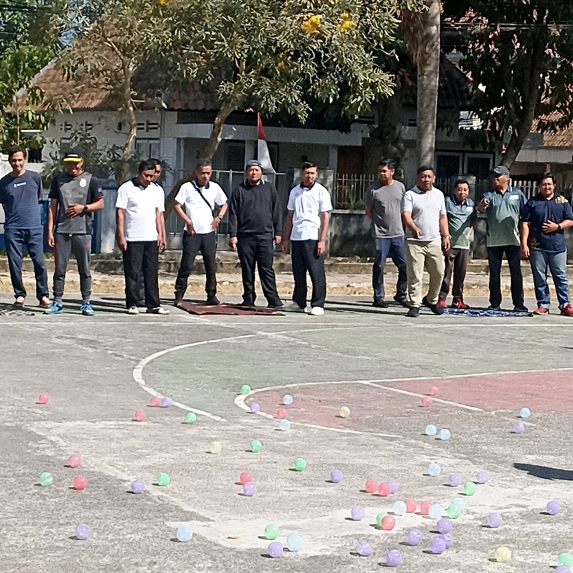 The image shows a group of approximately 15 men gathered on an outdoor basketball court in Magelang, Indonesia. The men are of various ages and are dressed in casual attire, with some wearing athletic wear and others in more formal shirts. Several men are standing in a line facing the camera, while others are observing. In the foreground, numerous colorful small balls are scattered across the court. Behind the men, there is a building with a tiled roof and some trees, suggesting an urban or semi-urban setting. A small Indonesian flag is visible on a flagpole in the background. The lighting suggests it is daytime, likely with bright sunlight casting shadows.