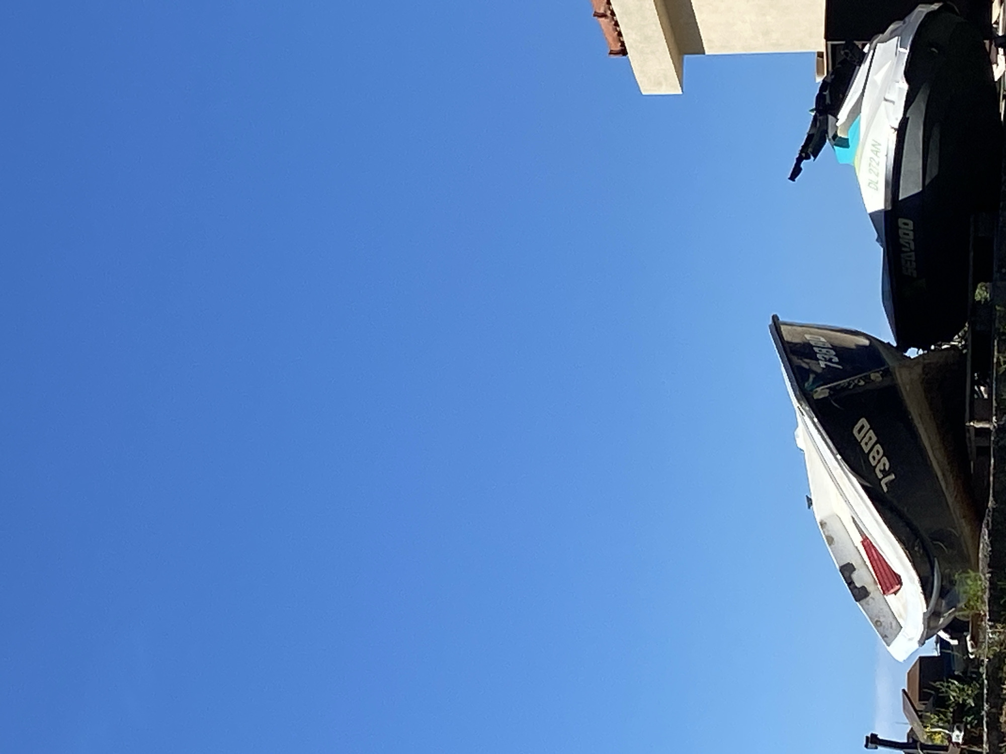 The image shows two watercraft on land under a clear blue sky. On the left is a dark-hulled boat with white trim, bearing the number "73800" on its side. A red object is visible inside the boat. To the right of this boat is a white and dark grey jet ski with "SEA-DOO" and "DL 272 AN" visible on its body. Behind the jet ski, a portion of a building with a terracotta-tiled roof is visible. The scene appears to be in Prijevor, Montenegro.