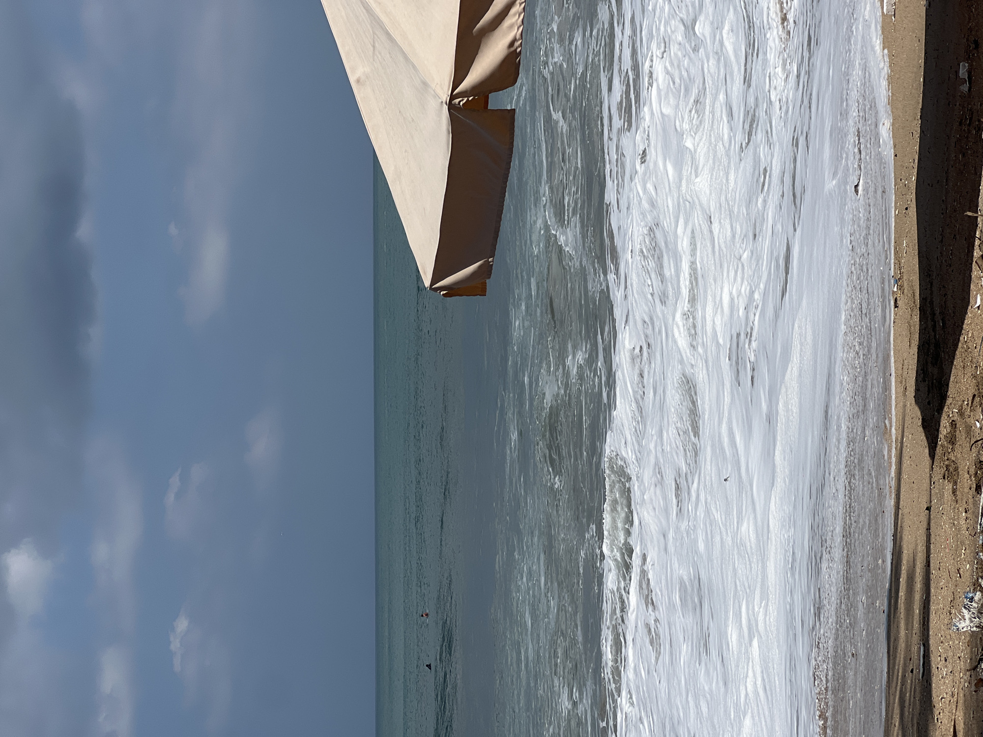 The image depicts a beach scene in Kuta, Indonesia. In the foreground, the sandy shore is visible with some debris scattered on it, and shadows cast by an unseen object. The middle ground is dominated by the ocean, with waves crashing and creating white foam as they reach the shore. A large, light-colored beach umbrella is positioned in the upper right portion of the frame, partially obscuring the view of the sky and sea. In the distance, on the calmer waters of the ocean, two figures can be seen, appearing to be in the water, possibly surfers. The sky above is a mix of blue and gray clouds, suggesting a partly cloudy day.