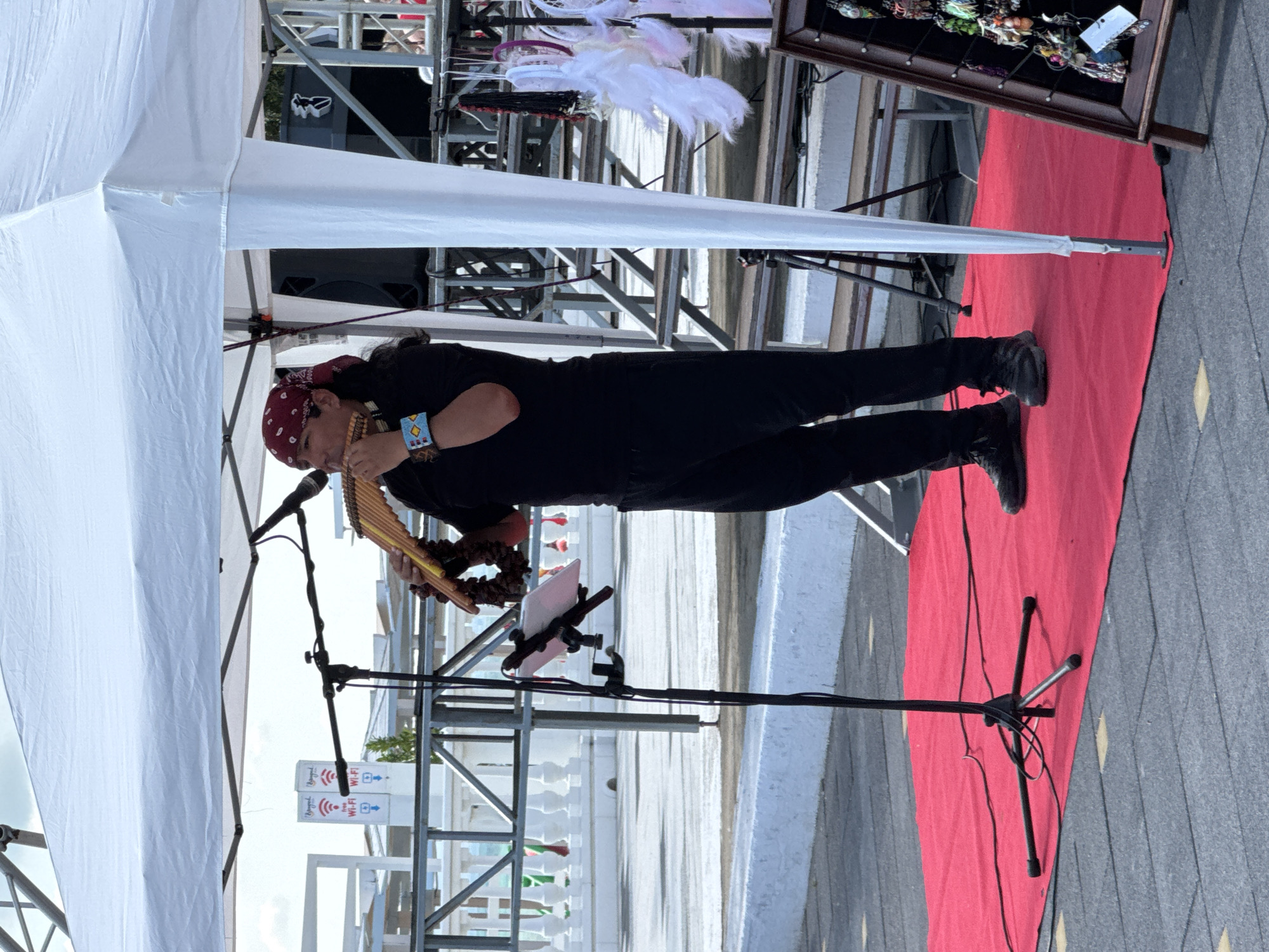 This image shows a person playing a pan flute under a white tent. The person is wearing a black t-shirt, black pants, and black shoes. They are also wearing a red bandana on their head and beaded bracelets on their wrists. The pan flute is made of wood and has multiple tubes of varying lengths. The person is standing on a red carpet, with a microphone stand to their left. In the background, there is a metal structure that appears to be part of a stage or display. There are also some items on display in a wooden case to the right of the person. Two signs with Wi-Fi symbols are visible on the left side of the image. The image appears to be taken outdoors during the daytime, with overcast weather. The overall scene suggests an outdoor performance or market event.