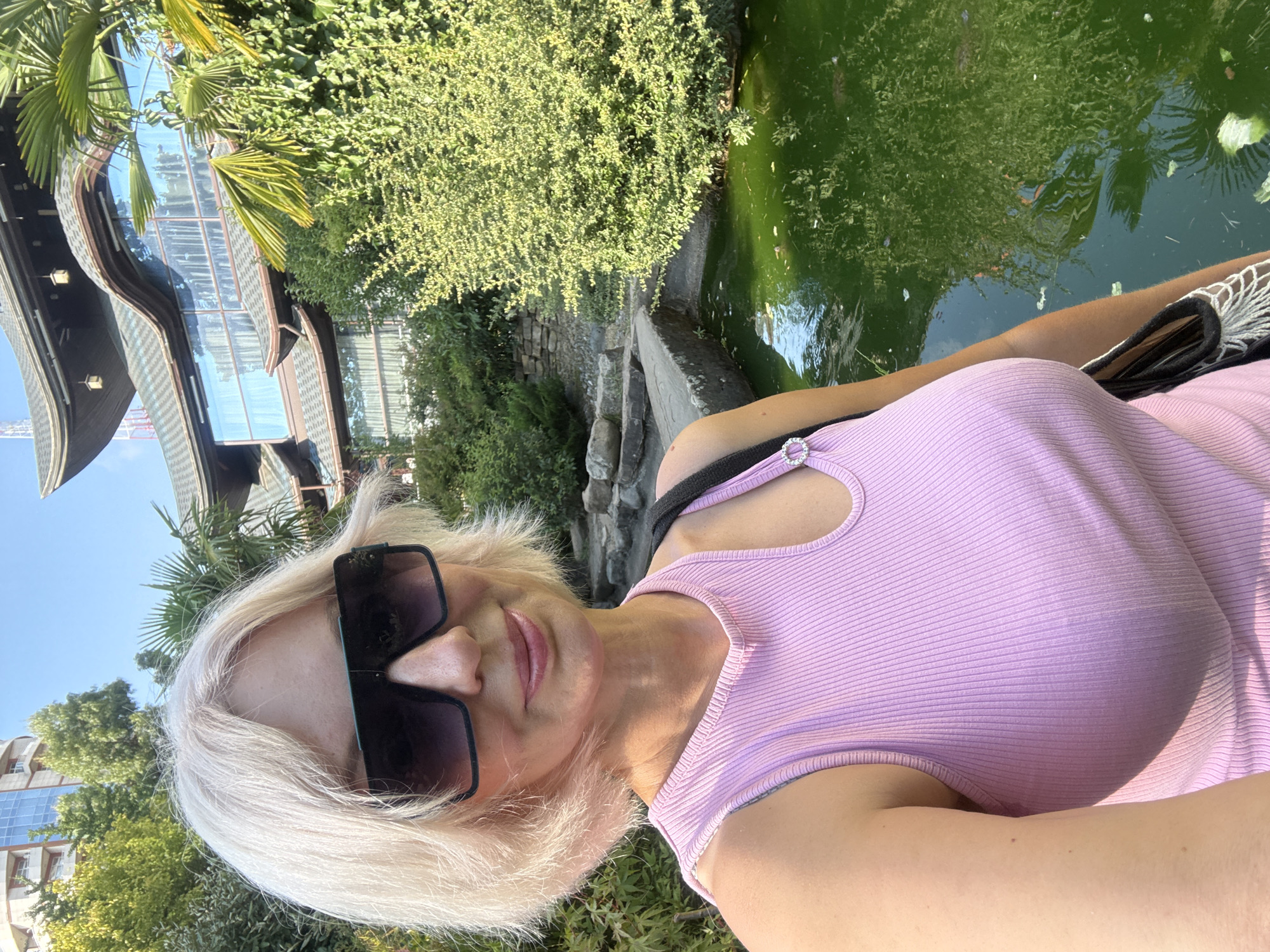 A fair-skinned woman with short blonde hair and large black sunglasses is taking a selfie outdoors. She is wearing a pink ribbed tank top with a cut-out detail at the shoulder and a rhinestone embellishment. She is standing in front of a body of green water, possibly a pond or a fountain, with lush green foliage and trees surrounding it. In the background, there is a building with a distinctive Asian-inspired architectural style, featuring curved roofs and large windows. This building appears to be part of a complex or resort. The sky is clear and blue, indicating a sunny day. The location is identified as the "Central Park of Culture and Recreation 'Riviera'" in the city of Sochi, Russia, known for its oriental-style architecture and landscaping.