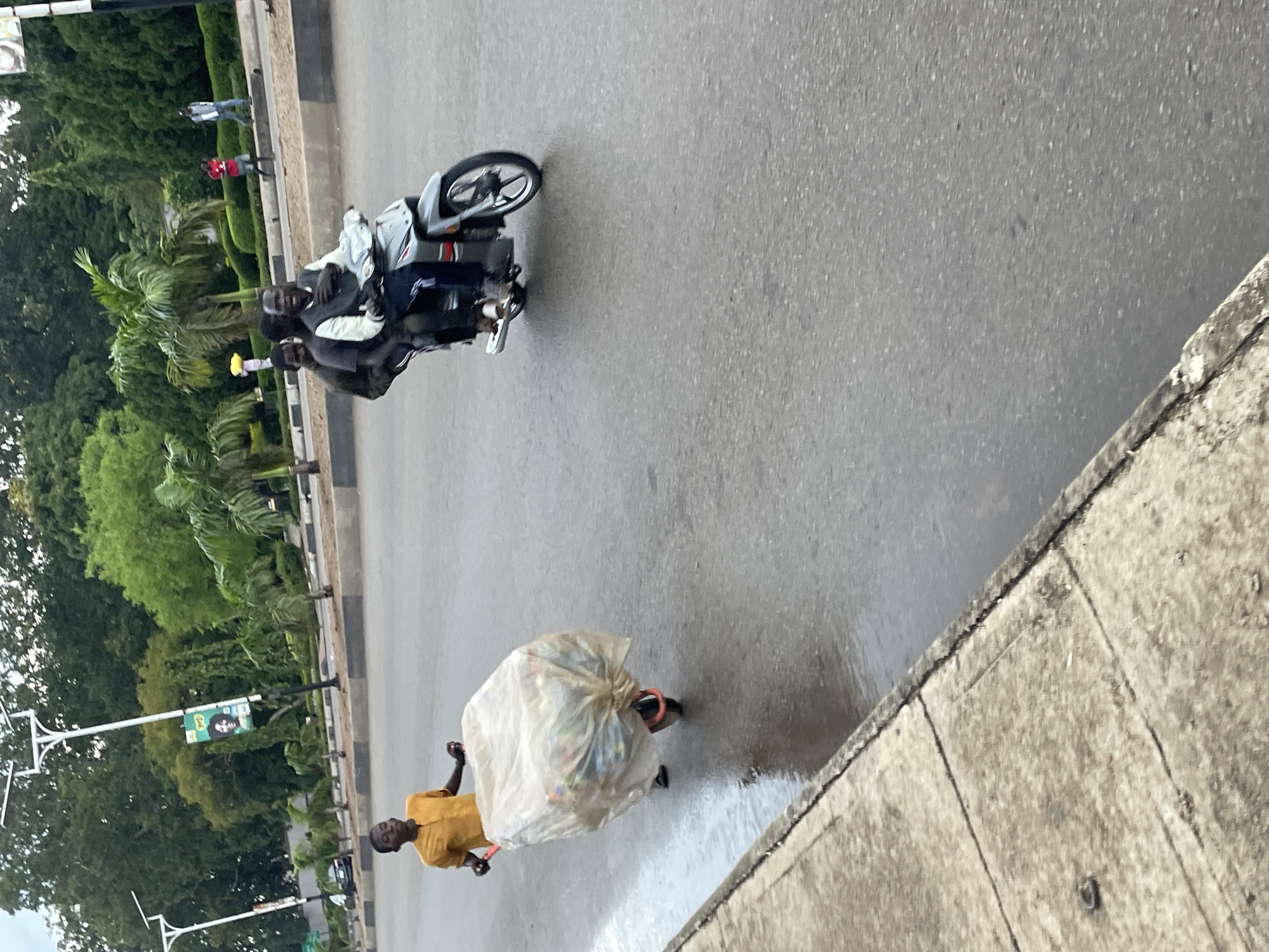 In Jos, Nigeria, a young person wearing a yellow shirt pushes a wheelbarrow overflowing with a large, transparent plastic bag filled with what appears to be recyclables. The wheelbarrow has a single wheel and red handles. The person is walking on a wet asphalt road, with a concrete sidewalk on the left. In the background, a motorcycle carrying three people is traveling in the same direction. The rider wears a dark jacket and blue pants, while the two passengers are seated behind him. Further in the background, lush green trees, palm trees, and streetlights line the road. A billboard with a picture of a person is visible on one of the lampposts. Two individuals, one in a red shirt and another in a light-colored shirt and dark pants, are walking on the sidewalk. The overall scene depicts a busy street in Jos, Nigeria, with everyday life activities taking place.