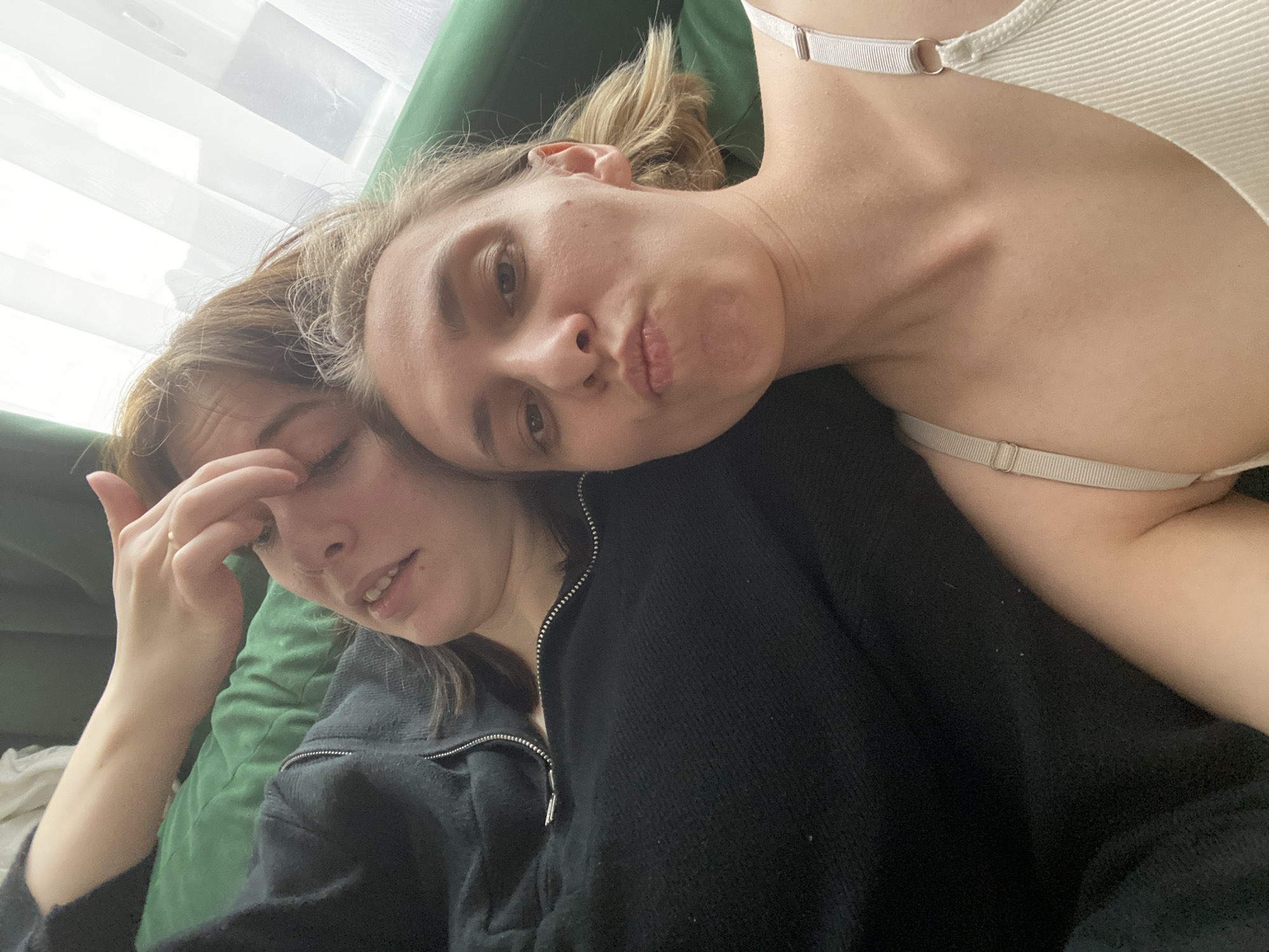 Two young women are lying on a green upholstered surface, possibly a sofa or bed. One woman, wearing a white tank top with visible bra straps, has her head resting on the other woman's shoulder. She is looking at the camera with slightly pursed lips. The other woman, wearing a dark zippered sweatshirt, has one hand touching her forehead or eye area. The background includes a white curtain or blind and part of the green upholstered surface. The location is indeterminate and cannot be identified as Chaikovsky, Russia, based solely on this image.