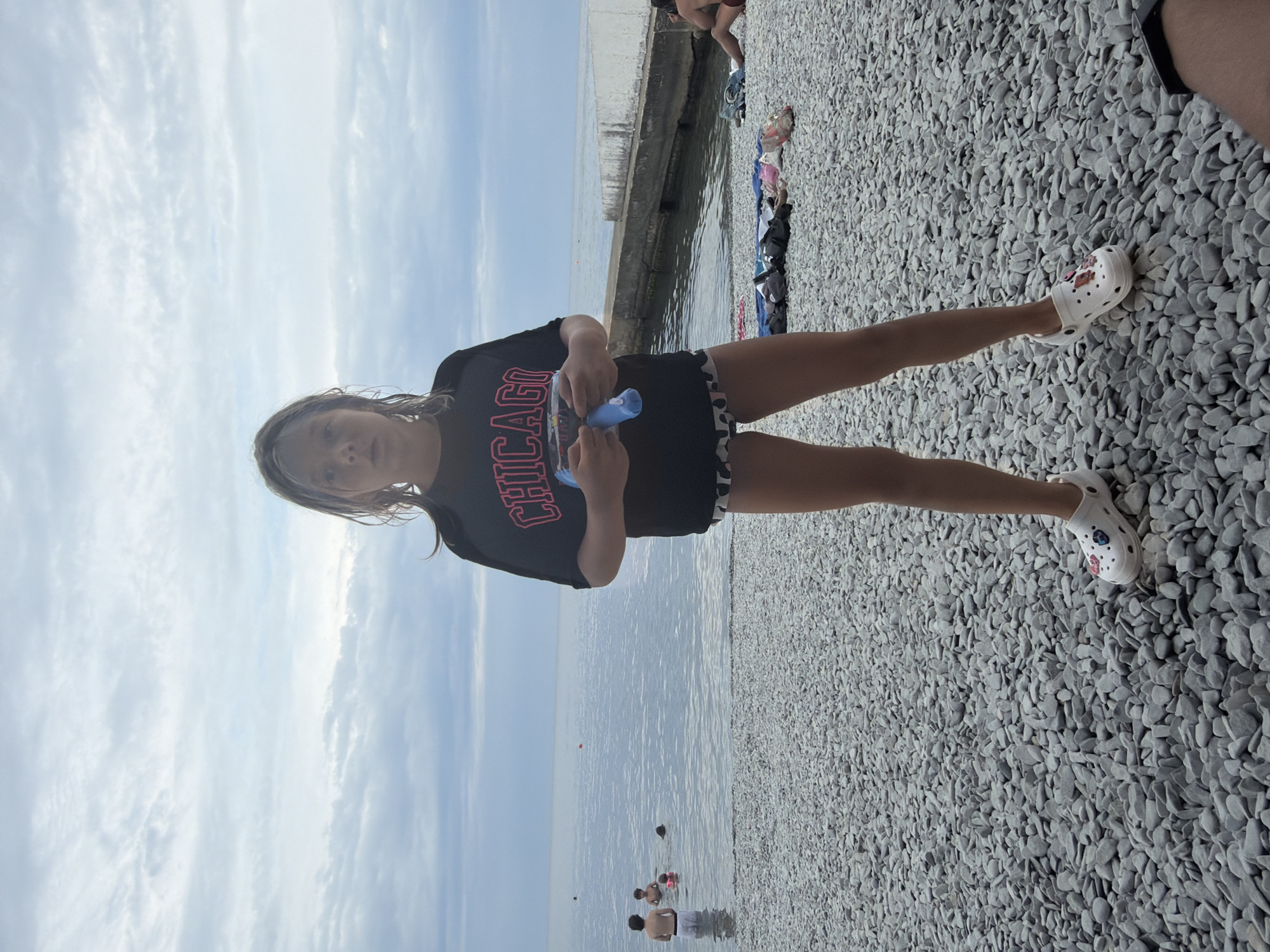 A full-shot, eye-level, outdoor photograph captures a young girl standing on a pebble beach, facing the camera. She is wearing a black t-shirt with "CHICAGO" in pink lettering, patterned shorts, and white Crocs. In her hands, she holds a blue and clear water gun. The beach is composed of small, grey pebbles, and in the background, the sea stretches to the horizon under a cloudy sky. Several people are visible in the water, some swimming and others wading. To the right of the frame, a concrete structure runs along the edge of the water. The location is the pebble beach in Partenit, Ukraine.