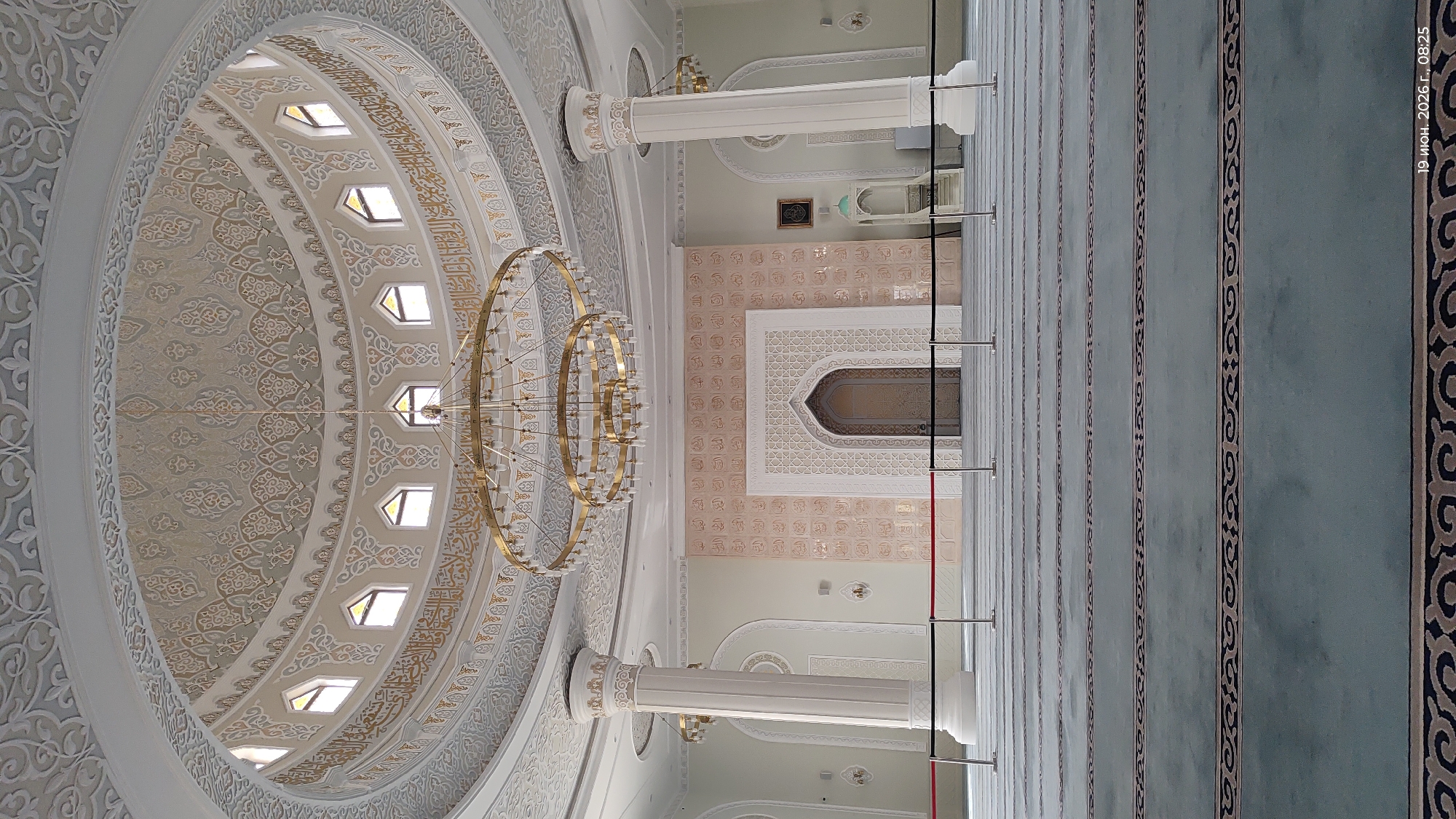The image depicts the interior of a large, ornate mosque, likely the Hazret Sultan Mosque in Astana, Kazakhstan, given its distinctive architectural features. The scene is empty of people, focusing instead on the grandeur of the space. The dominant colors are soft cream, beige, and pale green, with accents of gold and dark grey.
The perspective is from the floor, looking towards the front of the prayer hall. The floor is covered in a light green carpet with repeating dark grey geometric patterns, arranged in rows to guide worshippers. In the foreground, stanchions with red and black ropes create a temporary barrier, suggesting a controlled access or a demarcated area.
The walls are a pale cream color, adorned with intricate light beige decorative panels featuring Arabic calligraphy and geometric motifs. In the center of the far wall is a large, arched alcove with an ornate, gilded door. Above this alcove, the wall is decorated with repeating square panels, each containing calligraphy. To the right of this central feature, a dark framed artwork hangs on the wall, and a small, light blue minaret-like structure is visible.
Two massive, fluted columns, painted a creamy white with decorative gold bands near the top, flank the prayer hall. These columns support the magnificent domed ceiling. The dome is a masterpiece of Islamic architecture, featuring multiple layers of intricate stucco work in cream and grey, with golden Arabic inscriptions encircling its upper circumference. Small, arched windows are visible around the base of the dome, allowing natural light to enter.
A large, multi-tiered circular chandelier with a gold finish hangs prominently from the center of the dome, casting a warm glow. Smaller chandeliers are also visible on the sides of the main prayer hall. The overall impression is one of solemnity, elegance, and immense scale. The timestamp in the lower right corner indicates "19 June 2026, 08:25".