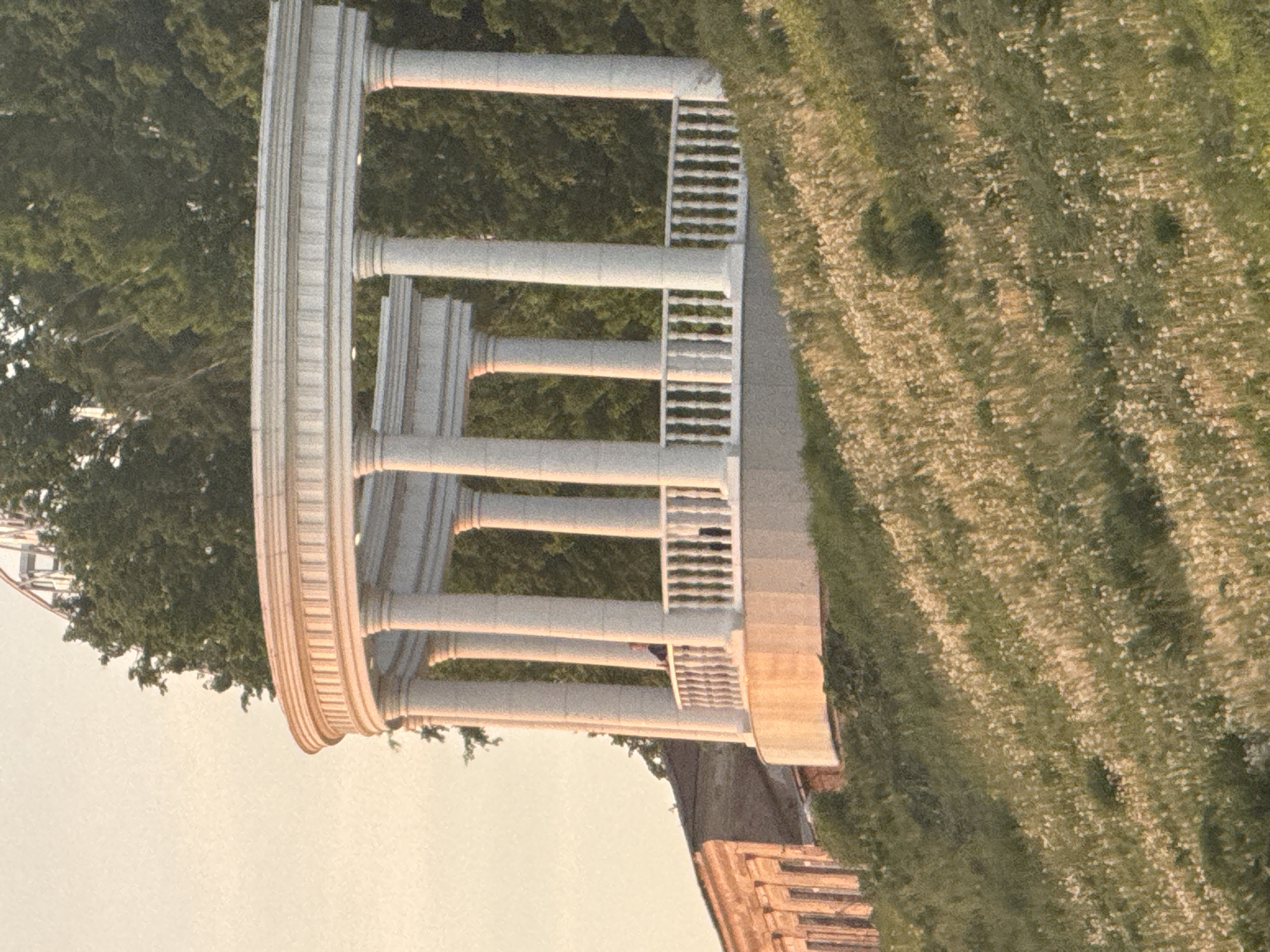 The image shows a rotunda structure with multiple pillars on a stone base, adjacent to a grassy area. There are trees in the background. Part of a ferris wheel is visible. The rotunda is in Izhevsk, Russia.