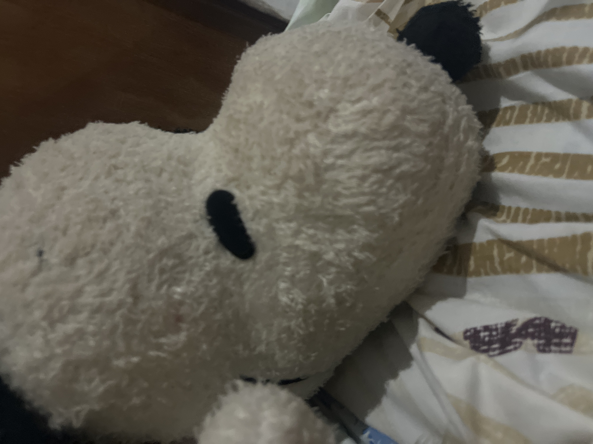 A close-up, slightly blurry, low-angle shot shows a stuffed toy, likely Snoopy, resting on a striped blanket. The toy is predominantly white and fluffy, with black markings for its eyes and ears. The blanket features alternating wide stripes of white and a light brown or tan color. A dark wooden surface, possibly furniture, is visible in the background on the right side of the image. The setting appears to be indoors, with the overall lighting suggesting it might be nighttime or a dimly lit room. There is no visible indication of the city of Santa Ana, Venezuela.