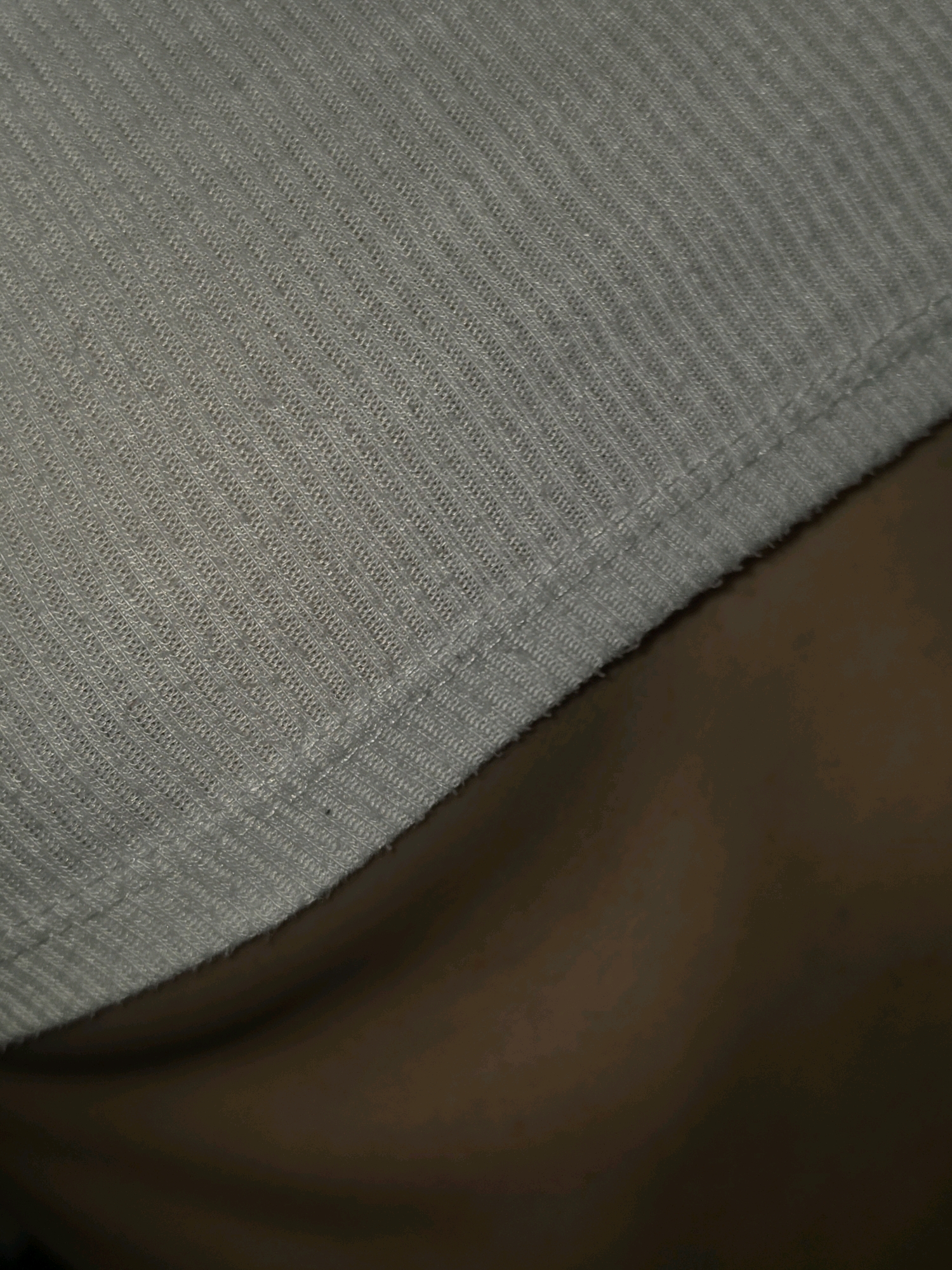The image displays a close-up view of a grey, ribbed fabric, possibly a garment. The fabric's texture is clearly visible, showing fine vertical ribbing. A subtle seam runs along the bottom edge of the fabric, and below it, a darker, smooth surface is visible, which appears to be skin. The lighting is soft, casting gentle shadows that highlight the texture of the fabric. The background is blurred and dark, suggesting a shallow depth of field. The scene is abstract and lacks any clear indication of a specific location within Pintomakmur, Indonesia, or any discernible actions or interactions.