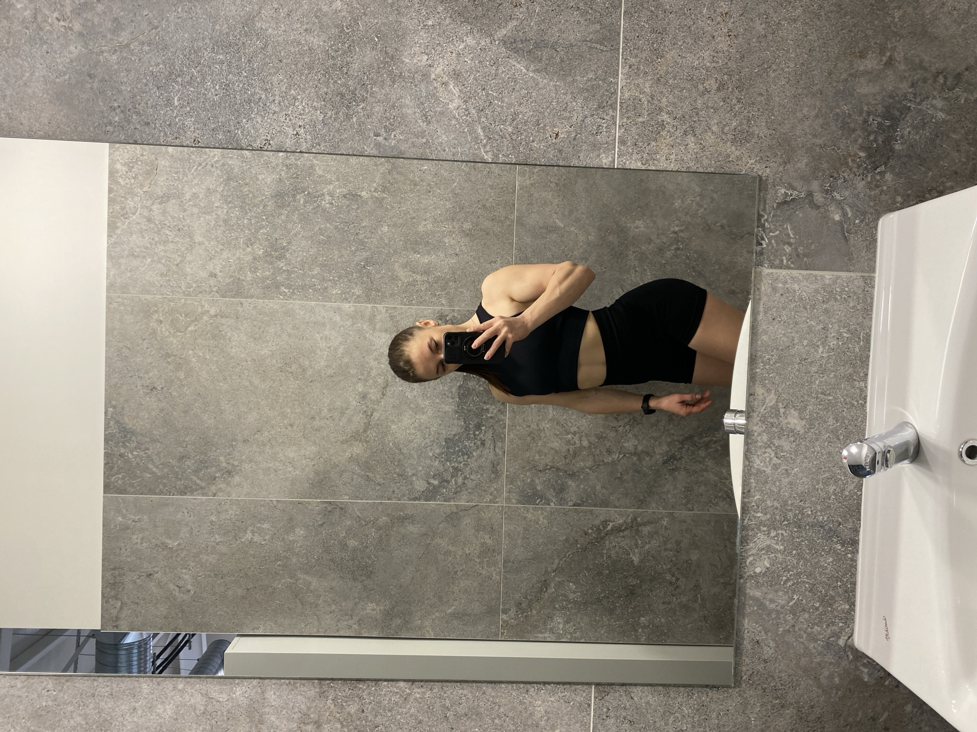 The image captures a mirror selfie taken by a person in what appears to be a modern bathroom or changing room. The individual, likely female, is reflected in a large rectangular mirror, posing while holding a smartphone to capture the image. She is dressed in black athletic wear, consisting of a crop top and shorts, and has her hair pulled back. A black watch is visible on her left wrist.
The background primarily features large, rectangular grey wall tiles with a textured, stone-like finish and visible grout lines. To the left of the mirror, a plain white vertical panel extends. On the right side of the image, a white ceramic sink with a chrome faucet is partially visible. Text on the sink indicates "Ideal Standard" and "Vidima," while "ECO" is discernible on the faucet. The bottom edge of the mirror reflects industrial-style overhead structures, such as ventilation ducts, suggesting the setting is a contemporary public facility, possibly a gym or sports center, located in Plovdiv, Bulgaria. The time of day and weather are not discernible from the image, and the person's expression is neutral, focused on taking the photo.