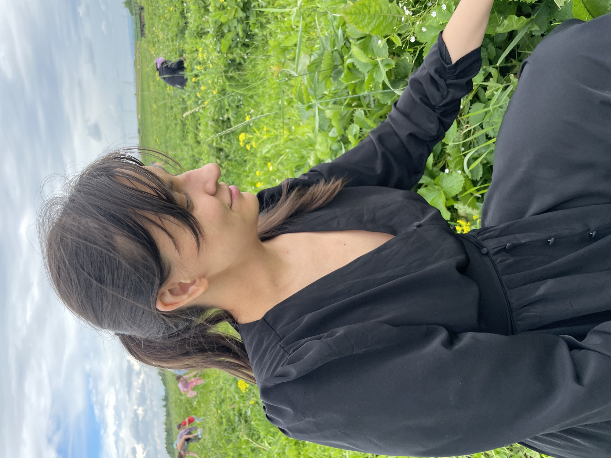 The image shows a young woman in a black dress, sitting in a field of green grass and yellow wildflowers under a cloudy sky. She is looking off to the side with a relaxed expression. In the background, other people, also seemingly dressed in casual clothing, can be seen bending over in the field, suggesting they are picking something from the ground. The overall impression is one of a pleasant, outdoor activity. The image appears to have been taken during the daytime.
