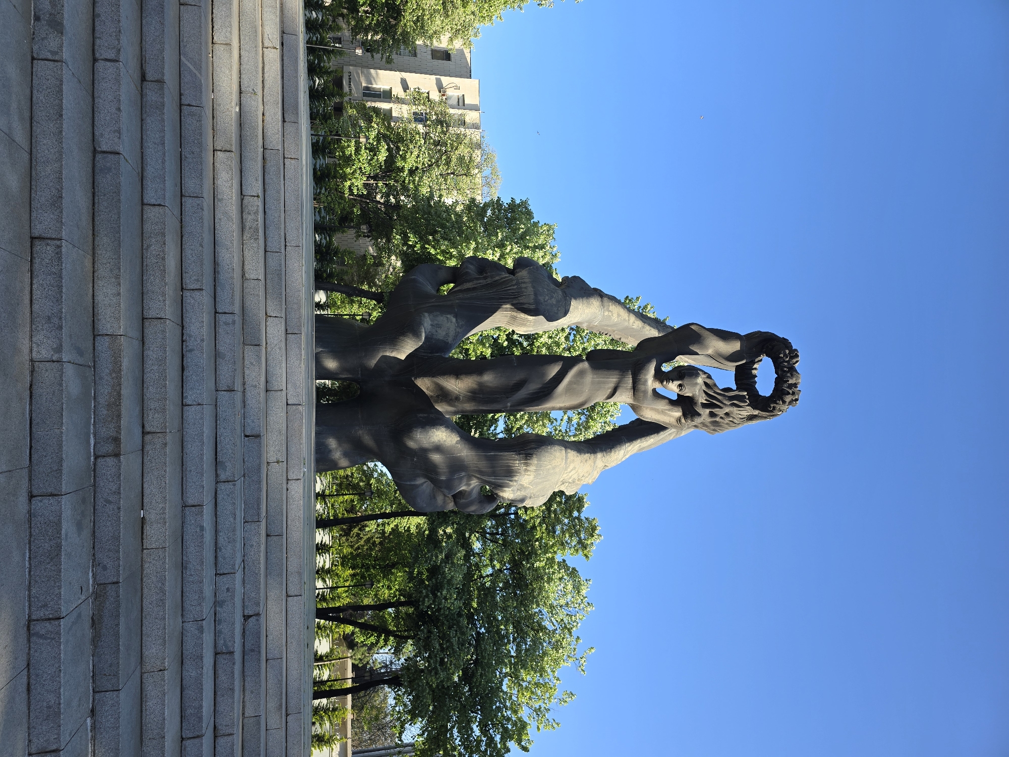 The image captures a statue in the city of Unknown, Unknown. The statue, made of a dark, possibly stone material, depicts figures holding up what appears to be a wreath or crown. The sculpture is set against a backdrop of trees and a clear blue sky. To the left of the sculpture are stone steps, leading up to the statue. The environment is urban, with a building visible in the background behind the trees.