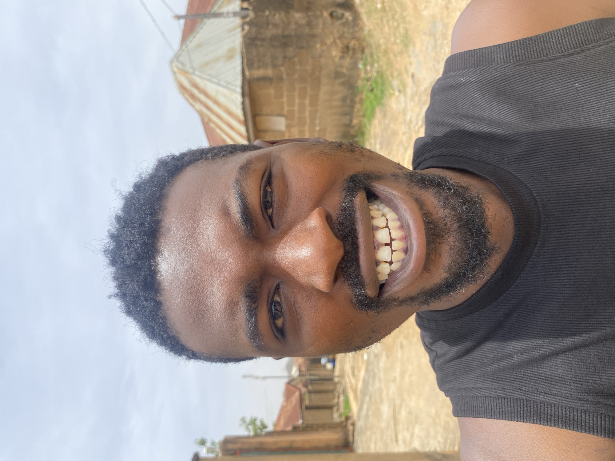 A close-up, portrait-style image captures a man with dark skin, smiling broadly and looking directly at the camera. He has short, curly black hair, a mustache, and a beard, and is wearing a black ribbed sleeveless shirt. His wide smile reveals his teeth, conveying a friendly and joyful emotion.
The background, slightly out of focus, indicates an outdoor setting during bright daylight, likely under an overcast or hazy sky. Visible structures include a building with a corrugated metal roof and another constructed from brick or mud brick, suggesting local architecture in a rural or semi-urban environment. The ground appears to be unpaved dirt or sand, with hints of sparse vegetation. The man's attire suggests a warm climate. The location is Barakin Akawo, Nigeria. There is no visible text in the image.
