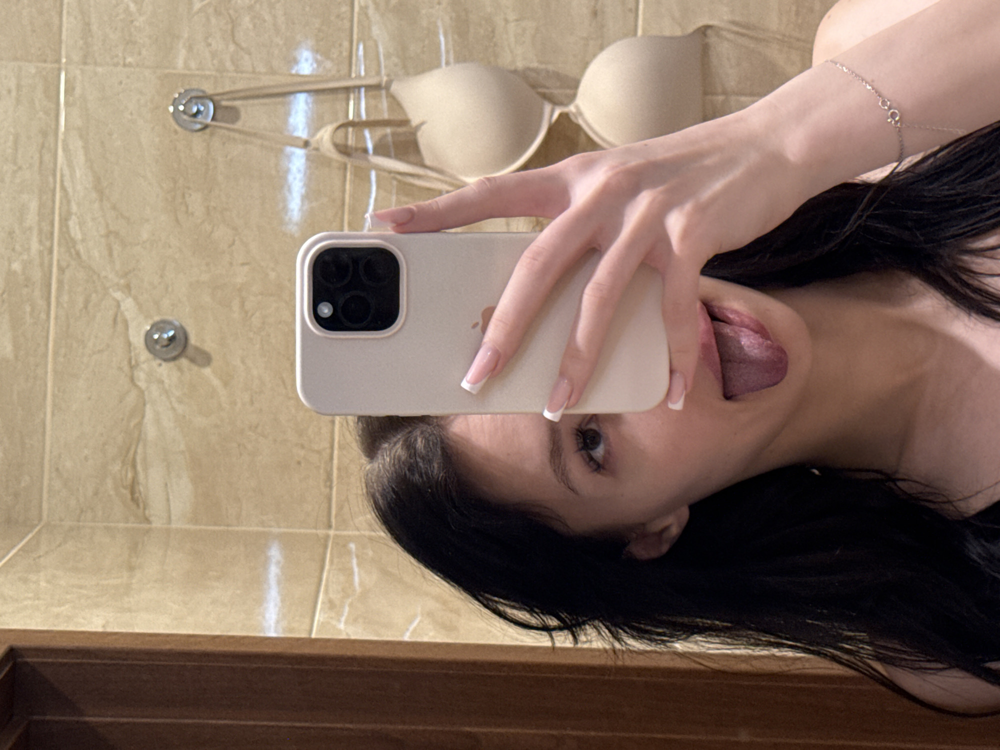 A young woman with long, dark hair is taking a selfie in a bathroom. She is holding a white iPhone in her right hand, covering part of her face. Her tongue is sticking out. A beige bra is hanging on hooks on the wall behind her. The walls are tiled in a beige, marble-like pattern. A wooden doorframe is visible on the left side of the image. A thin bracelet with a small charm is visible on her left wrist. The lighting suggests an indoor environment, likely a home bathroom. The image does not provide any specific indicators of the city of Khimki, Russia.