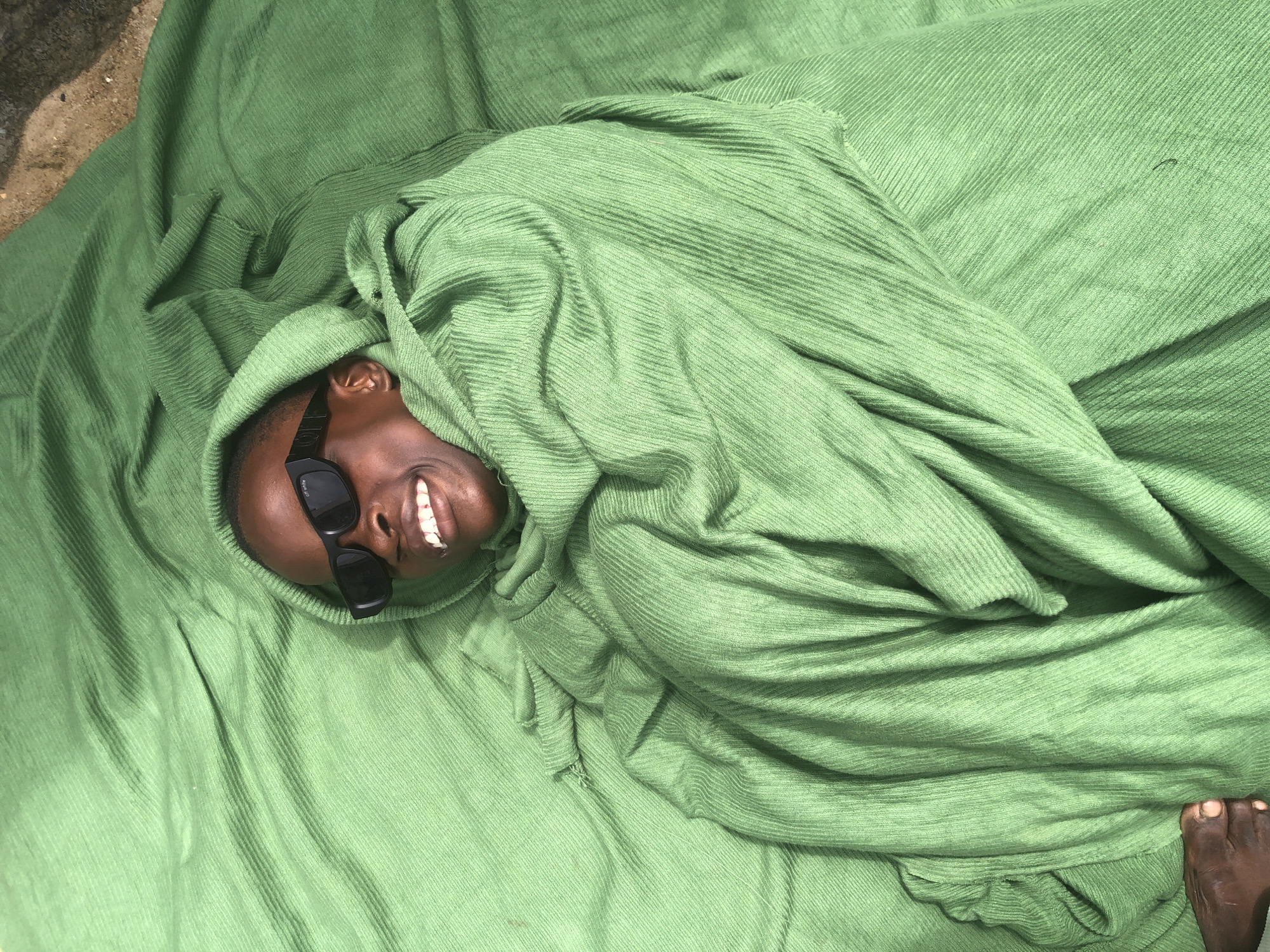 A young person wearing black sunglasses and a green hooded garment is lying down on a green fabric that is draped over them. The person is smiling, with their teeth visible. Their head is turned to their right, and their face is illuminated by natural light. A portion of their foot is visible in the lower left corner of the image. The fabric appears to be a ribbed material. The background is out of focus but suggests an outdoor setting with a textured surface, possibly dirt or sand. The overall impression is one of relaxation and comfort. This scene is situated in Ado, Nigeria.