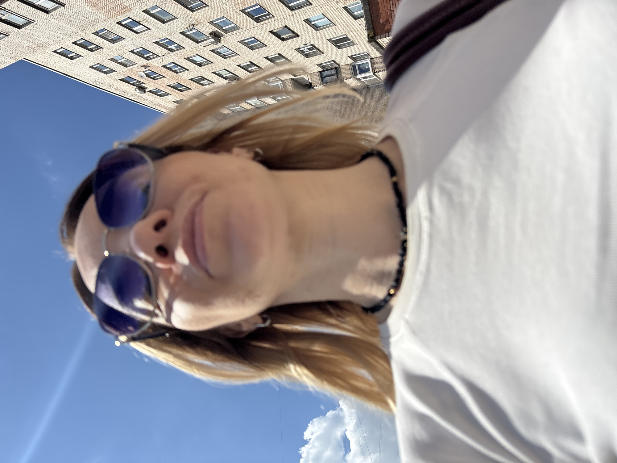 The image is a vertical close-up, rotated 90 degrees counter-clockwise, presenting a low-angle shot of a person outdoors. The main subject is a person, appearing female, with blonde hair that is slightly wind-blown. She wears dark-rimmed sunglasses with a blue tint, concealing her eyes, and her mouth shows a subtle, relaxed smile. A dark, possibly beaded, necklace is visible around her neck, and she is dressed in a white t-shirt or top, with a dark band or stripe visible on the right shoulder.
The background features a clear, bright blue sky with a few white, fluffy clouds visible towards the lower right portion of the frame (when the image is viewed with the person upright). Above the sky, a large, multi-story brick building dominates the upper background. This building has a facade of light-colored bricks and numerous regularly spaced rectangular windows, some of which appear to be dormers or skylights, suggesting an urban architectural style.
The activity captured is a casual outdoor moment, likely a selfie taken from a low perspective or a photograph where the subject is looking slightly away from the lens. The bright sunlight indicates it is daytime, under sunny and clear weather conditions. The overall setting is an urban environment, and the provided context places this scene in Saint Petersburg, Russia. There is no visible text in the image.