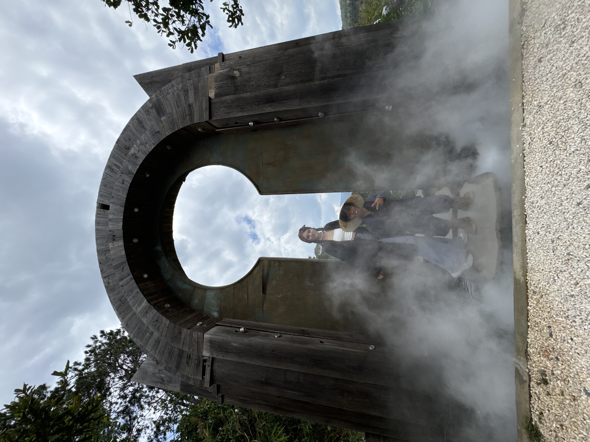 Two people are standing in a steaming wooden archway in what appears to be Thôn 1, Vietnam. The archway is made of rough-hewn wooden planks and has a circular opening at the top. Steam is billowing from the area where the people are standing. One person is wearing a hat, and the other has a bandana in their hair. There is vegetation visible both above and below the archway. The sky is overcast.