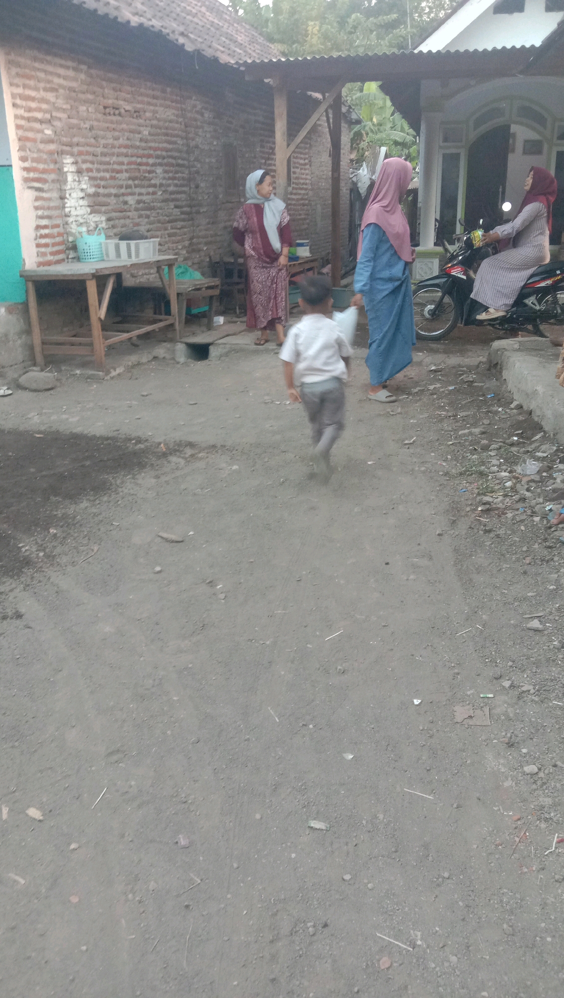 The image depicts an outdoor scene in Karang Anyar, Indonesia, likely a residential area. In the foreground, a young child wearing a white shirt and grey pants is running away from the camera. Behind the child, three women are visible. The woman on the left wears a patterned maroon dress and a light grey hijab. Next to her, a woman in a blue dress and a pink hijab is walking. Further to the right, a woman in a striped dress and a maroon hijab is seated on a motorcycle. The environment includes a brick building with a corrugated metal roof, a wooden table with baskets, a smaller wooden stool, and a white house with a tiled roof. The ground is a dirt or gravel pathway with some scattered debris. The lighting suggests it is daytime, possibly late afternoon.