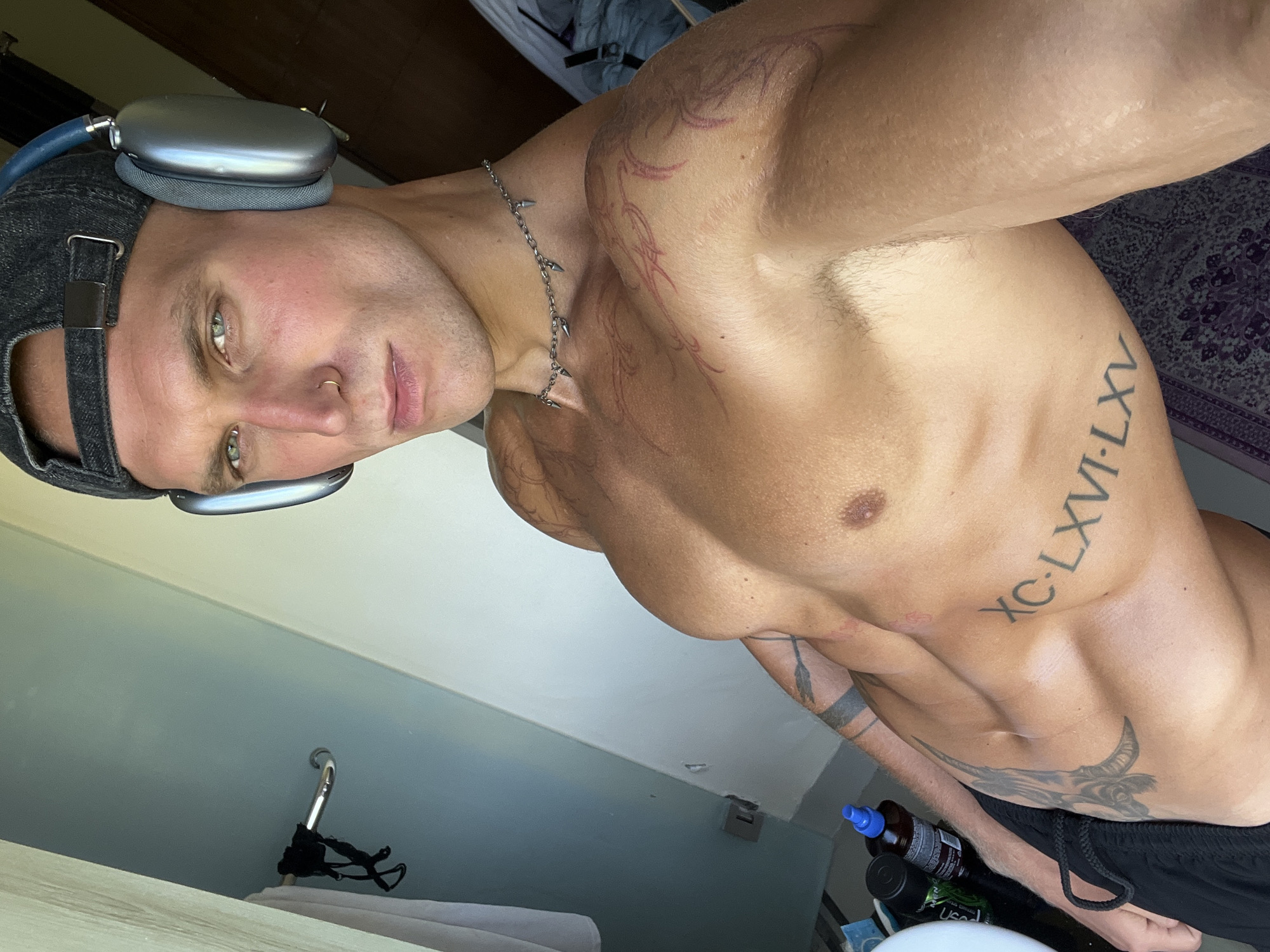 A man with blue eyes, a nose ring, and tattoos on his chest and arms, wearing a dark baseball cap and silver headphones, poses for a selfie in what appears to be a hotel room in North Kuta, Indonesia. His torso is bare, revealing muscular abs and pectoral muscles. He is wearing black shorts. In the background, a light-colored wall and a wooden door are visible. A towel hangs on a hook on the wall. On a surface to the left of the frame, there are several bottles, possibly toiletries or supplements. The lighting suggests it is daytime, with light coming from the right side of the image.