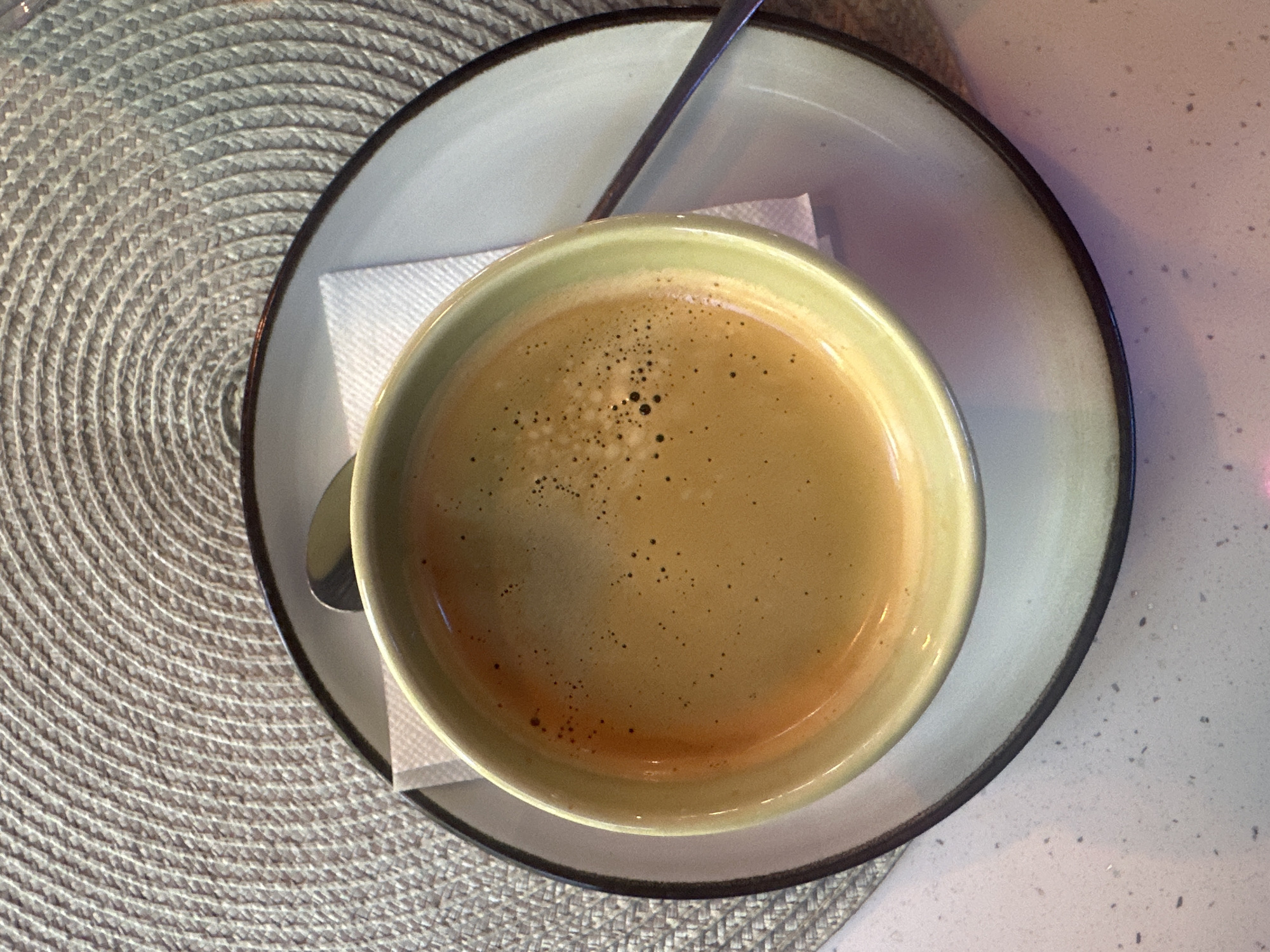 A close-up, overhead shot captures a light green ceramic cup filled with a dark liquid, likely coffee, resting on a white plate. The cup is positioned on a textured, circular placemat with a woven pattern. A white napkin is visible beneath the cup. A silver spoon lies on the plate, its handle extending to the right. The scene is set on a light-colored countertop with speckled dark flecks. The image's lighting creates subtle shadows and highlights, particularly on the surface of the coffee, revealing a light crema with small bubbles. The overall impression is one of a simple, neatly arranged beverage setting. The image does not contain any information or visual cues to indicate its location in Plesetsk, Russia.