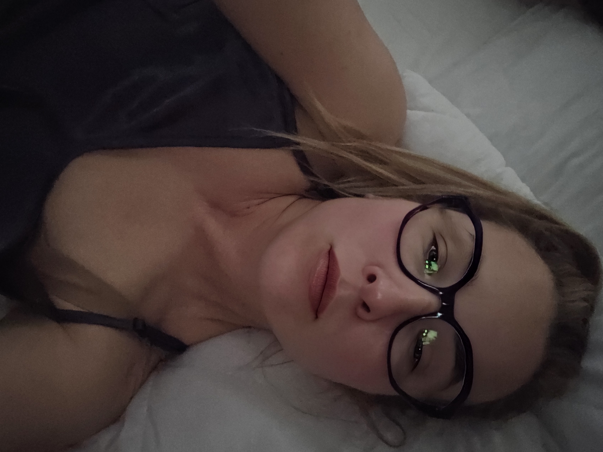A woman with light skin and light brown hair, wearing glasses with dark frames, is lying on her back on a white pillow and what appears to be a white sheet. She is wearing a dark-colored tank top with thin straps. The background is blurry and indistinct, suggesting an indoor setting. The image does not provide any information to suggest a location in Moscow, Russia.
