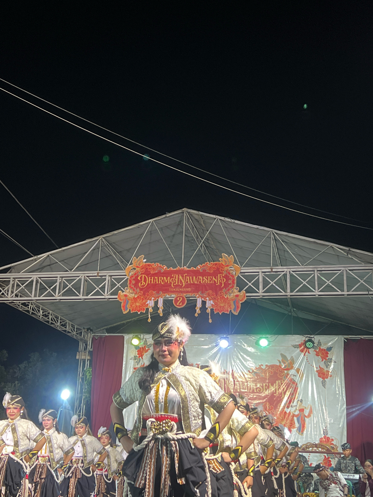The image shows a group of dancers in traditional attire performing on a stage at night in Madyocondro, Indonesia. The dancers, predominantly female, are wearing ornate costumes with white, gold, and black elements, including feathered headdresses and intricately patterned skirts. The main dancer in the foreground is a woman with elaborate makeup, adorned in a white and gold top, a black skirt with layered, patterned fabric, and a white feathered headdress. She is looking towards the camera with a serious expression. Behind her, several other dancers in similar costumes are arranged in a line, some with their hands clasped. The stage is set up under a large, open-air tent structure with metal scaffolding. A red banner with gold lettering that reads "DHARMA NAWASENA" and "TANDUNGSARI" is prominently displayed above the stage, along with the number "2". The backdrop of the stage features a white cloth with floral and avian motifs in red and gold. Several stage lights, some colored green and blue, illuminate the performers. In the background, a keyboardist and another individual are visible. The overall scene suggests a cultural performance or festival taking place outdoors at night.