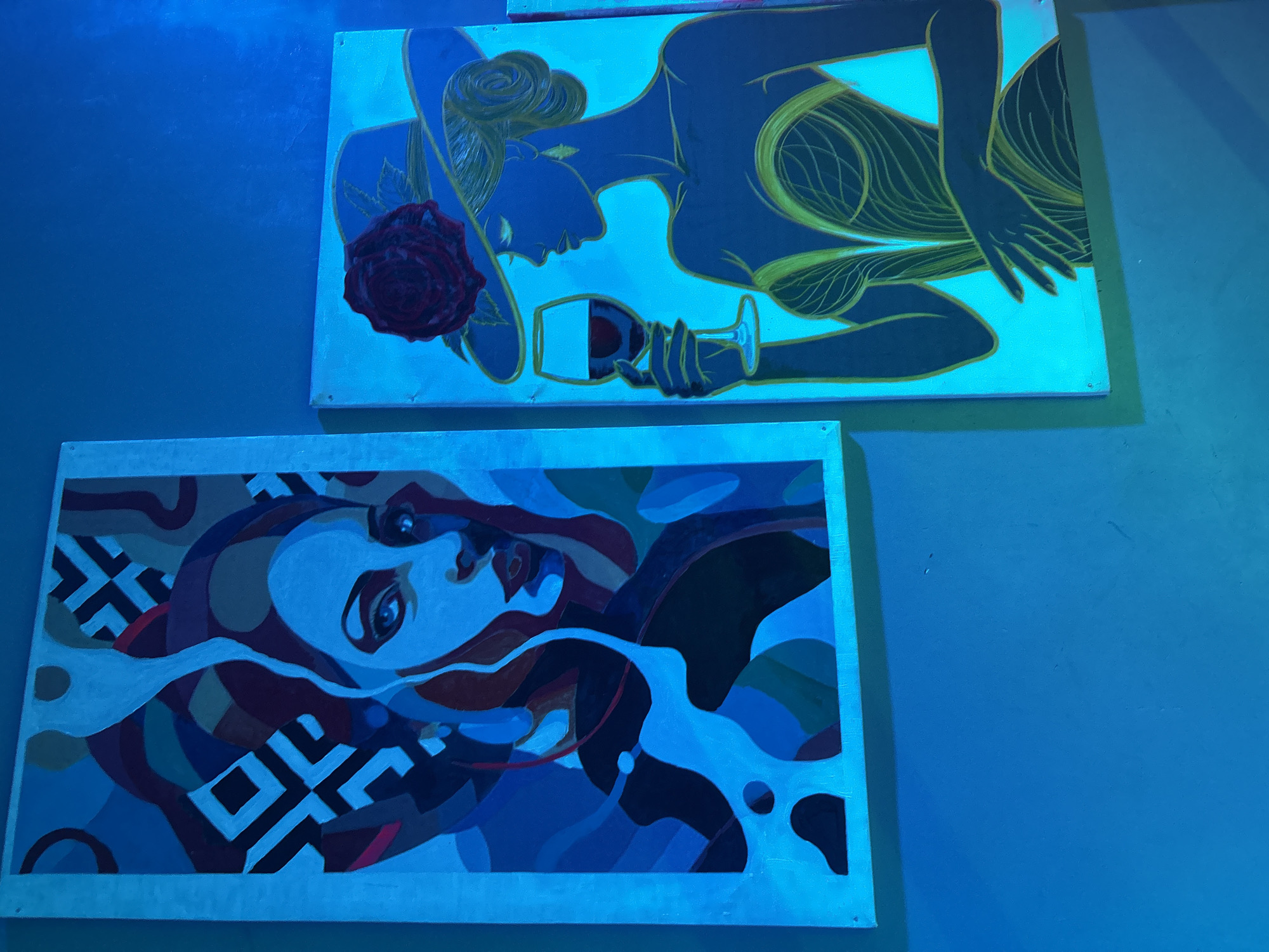 Two framed paintings hang on a blue wall. The top painting depicts a woman with a hat and a rose, holding a wine glass. The lower painting is an abstract portrait of a woman's face. There is no information in the image to determine its location in Lafia, Nigeria.