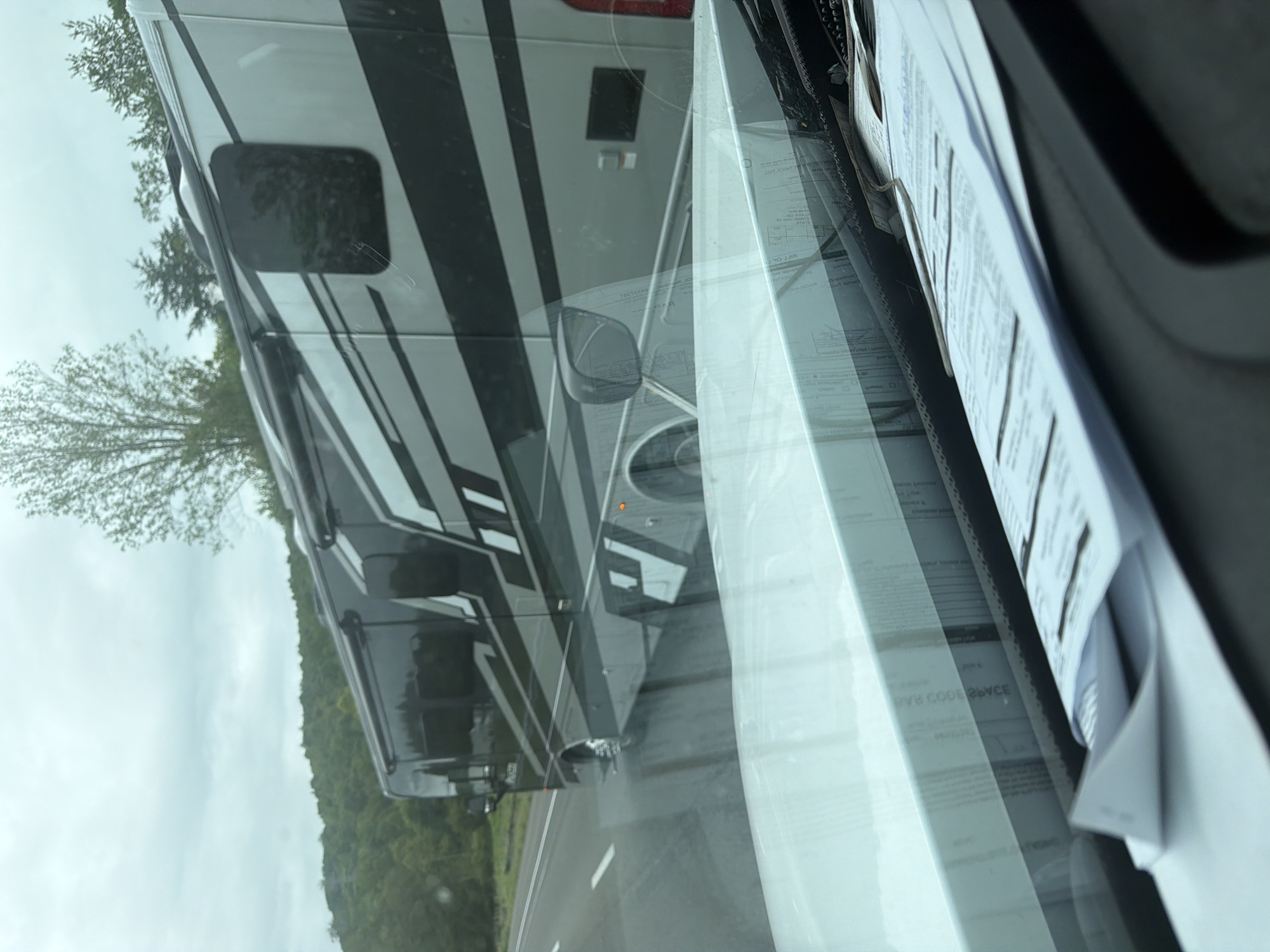 The image shows a side view of a large recreational vehicle (RV) driving on a highway. The RV is silver and black with a striped design. It has several windows and is traveling on the right side of the road. The environment appears to be rural or suburban, with trees and rolling hills visible in the background. The sky is overcast. In the foreground, the dashboard of the vehicle from which the photo is taken is visible, with papers and documents scattered on it. There are no people visible in the image. The scene is captured from a moving vehicle, as indicated by the slight blur and the perspective.