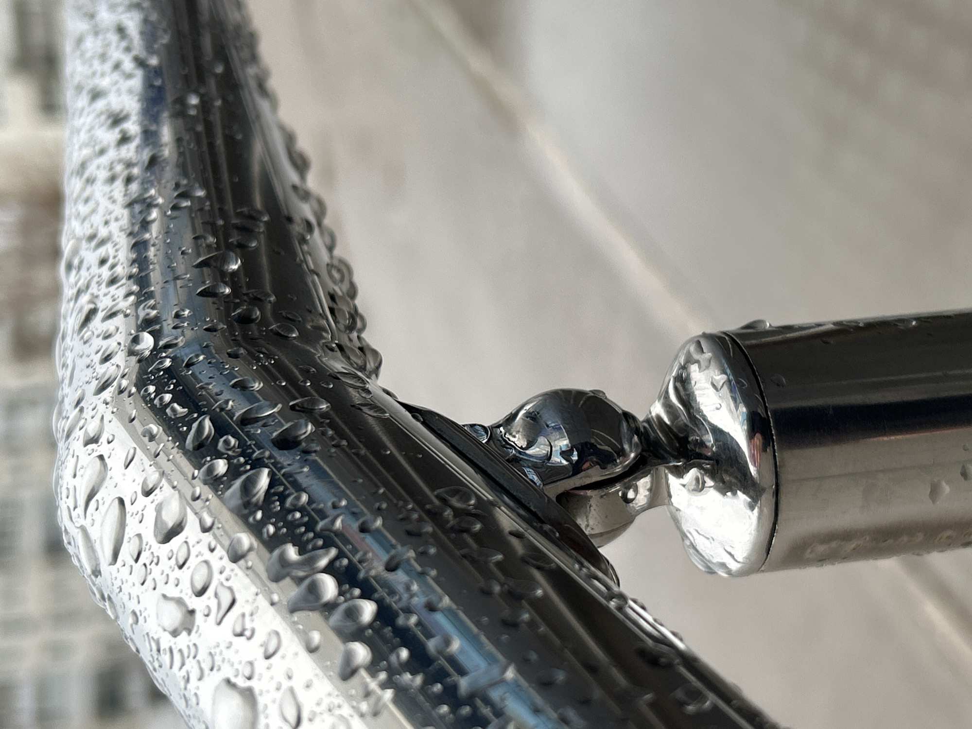 This close-up photograph captures a section of a shiny, metallic railing or pipe, heavily adorned with numerous water droplets. The metal, likely polished stainless steel or chrome, exhibits strong reflections, which are distorted by the varied shapes and sizes of the water beads clinging to its surface. The railing makes a distinct bend or features a joint in the middle-right of the frame, where two sections connect via a rounded, highly reflective fitting, also wet with droplets.
The lighting suggests it is daytime, and the presence of the water droplets strongly indicates that it has recently rained or condensation has formed. The focus is sharp on the metal and water, with a shallow depth of field rendering the background as a soft, out-of-focus blur of light grey and white tones. While indistinguishable, the blurred background might represent an exterior wall, another structure, or an overcast sky. The image conveys a sense of stillness and highlights the textures of water against a smooth, reflective surface. No people or visible text are present in the image, set in Chaikovsky, Russia.