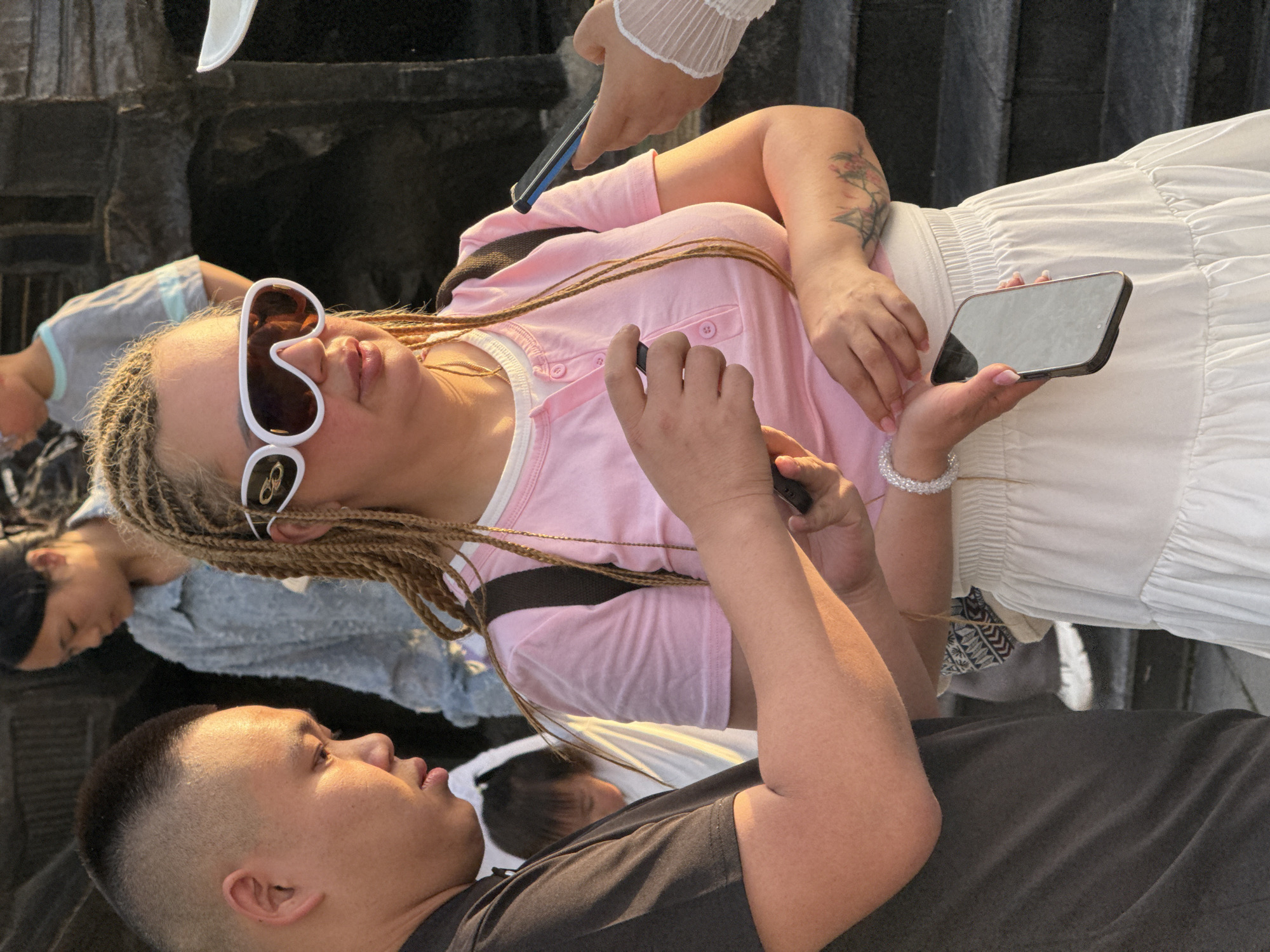 The image depicts a scene in Yuzhong District, China, where a group of people are interacting near what appears to be a reflective surface. The main focus is a woman with blonde braided hair wearing oversized white sunglasses, a pink shirt, and a white skirt. She is holding a smartphone. Another person is taking a photo of her with a different phone. A man is visible in the foreground wearing a black shirt and facing the woman. Additional people, including children, are reflected in the surface, indicating they are part of the group. The environment appears to be urban.