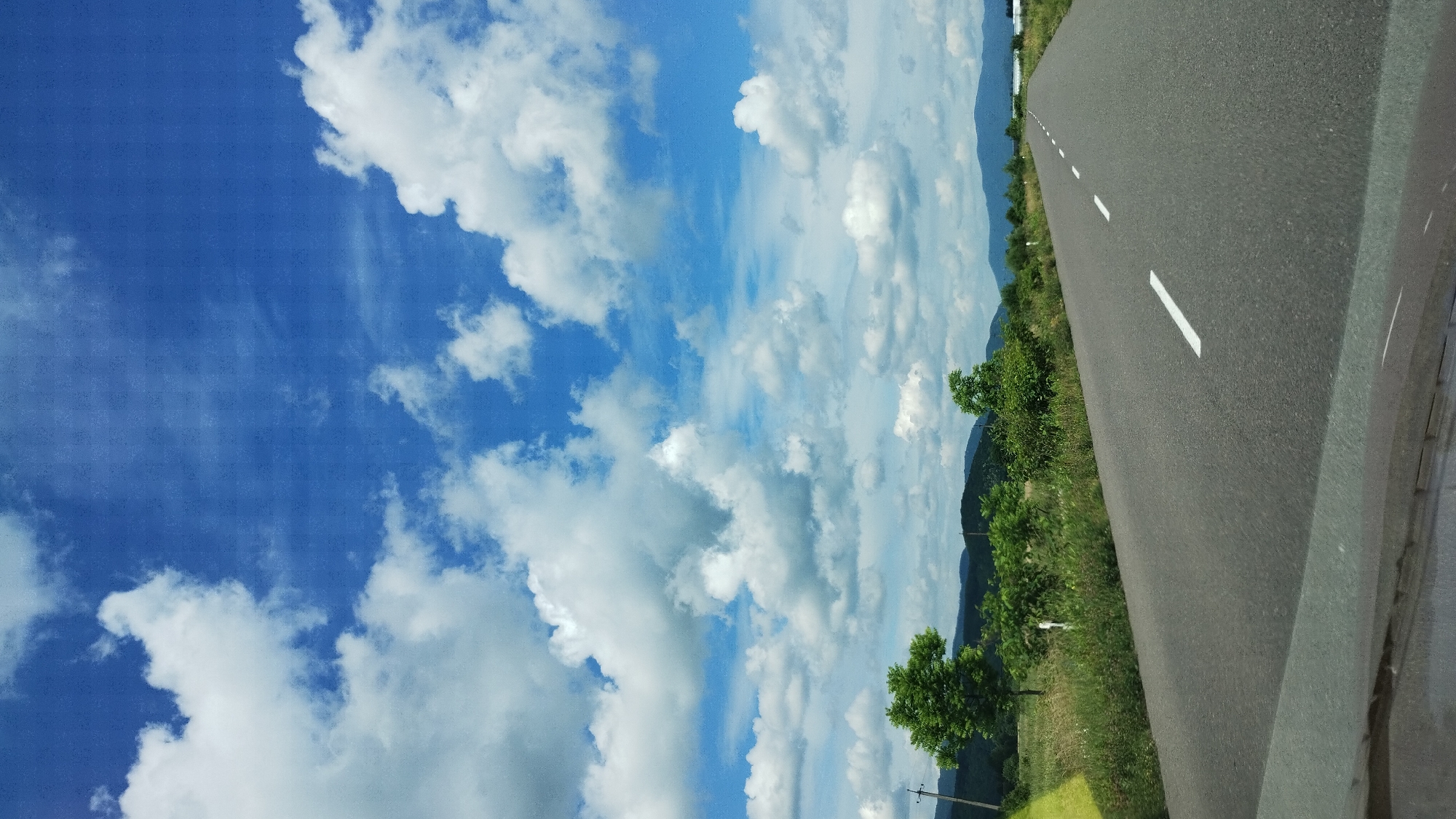 The image depicts an asphalt road stretching into the distance, bordered by lush green vegetation and rolling hills under a bright blue sky filled with fluffy white clouds. The road has a dashed white line marking its center. The landscape appears to be rural, with scattered trees and fields visible along the sides of the road. In the distance, mountains can be seen. The photograph seems to be taken from a vehicle traveling on the road. The location is Velyatyno, Ukraine.