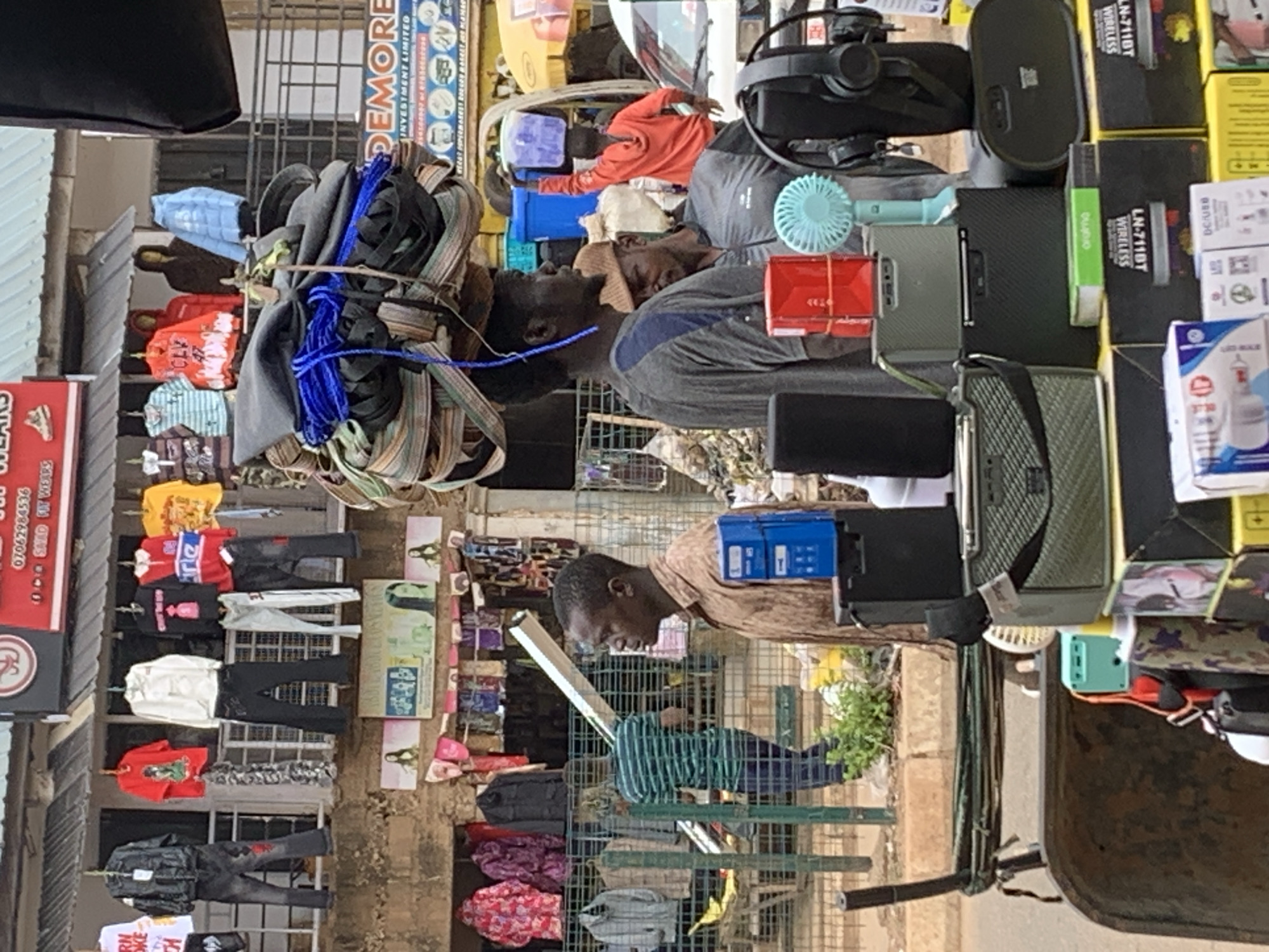 This image captures a bustling outdoor market scene in Bukuru, Nigeria. The primary subjects are two men, one leaning forward with his head bowed and the other wearing a red shirt and leaning back. They appear to be either vendors or shoppers in a crowded environment.
The scene is filled with various objects typically found in a market. Piles of clothing, including t-shirts and pants, are displayed, some hanging on racks. Various electronic items are also visible, such as headphones, a portable fan, what appear to be speakers, and boxes with labels like "WIRELESS" and "LN-7118T". There are also some plastic crates and bundles of rope or fabric.
A prominent sign with the text "DEMORE INVESTMENT LIMITED" and a phone number "07062984536" provides a location cue. The presence of multiple stalls and the density of people suggest a busy market day. The lighting indicates it is likely daytime. The overall atmosphere appears to be active and perhaps a bit chaotic, typical of a lively marketplace. The emotion of the men isn't clearly discernible due to the image quality and angle, but the man in the red shirt seems relaxed, while the other is more engaged with something unseen. The setting is clearly an open-air market with a somewhat rustic infrastructure, including metal shelving and awnings.